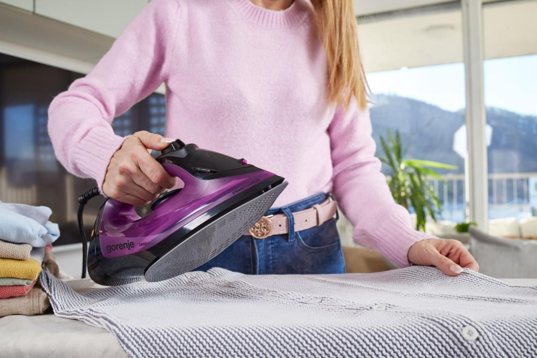 Osoba prasuje pasiastą koszulę fioletowym żelazkiem.