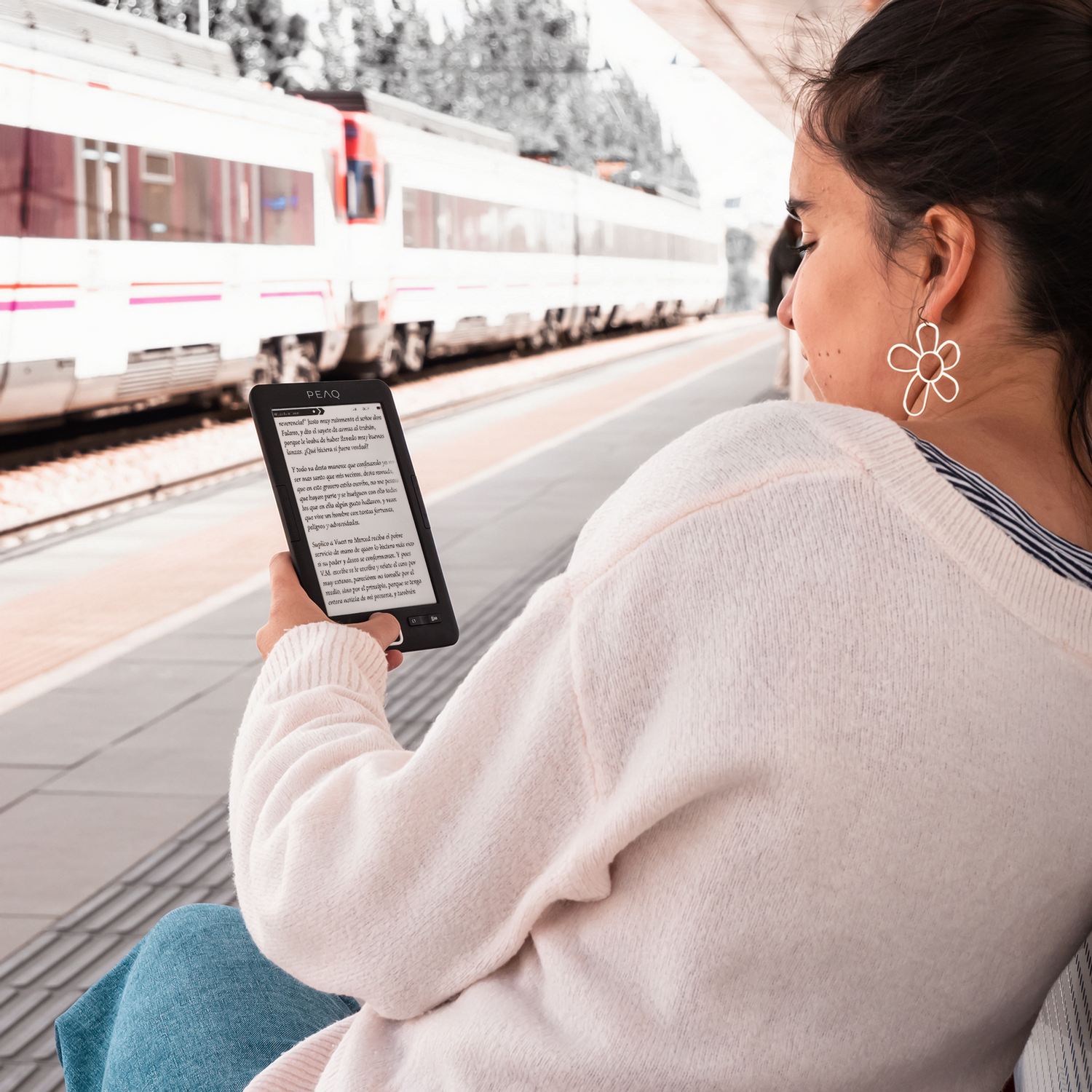 Una joven lee un libro electrónico en un andén mientras pasa un tren al fondo.