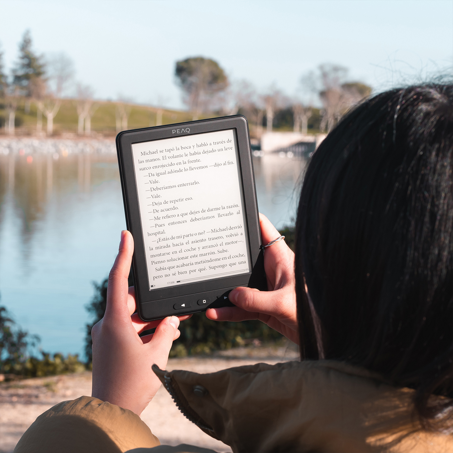 Persona sosteniendo e-reader con contenido de libro al aire libre, lago y árboles detrás.