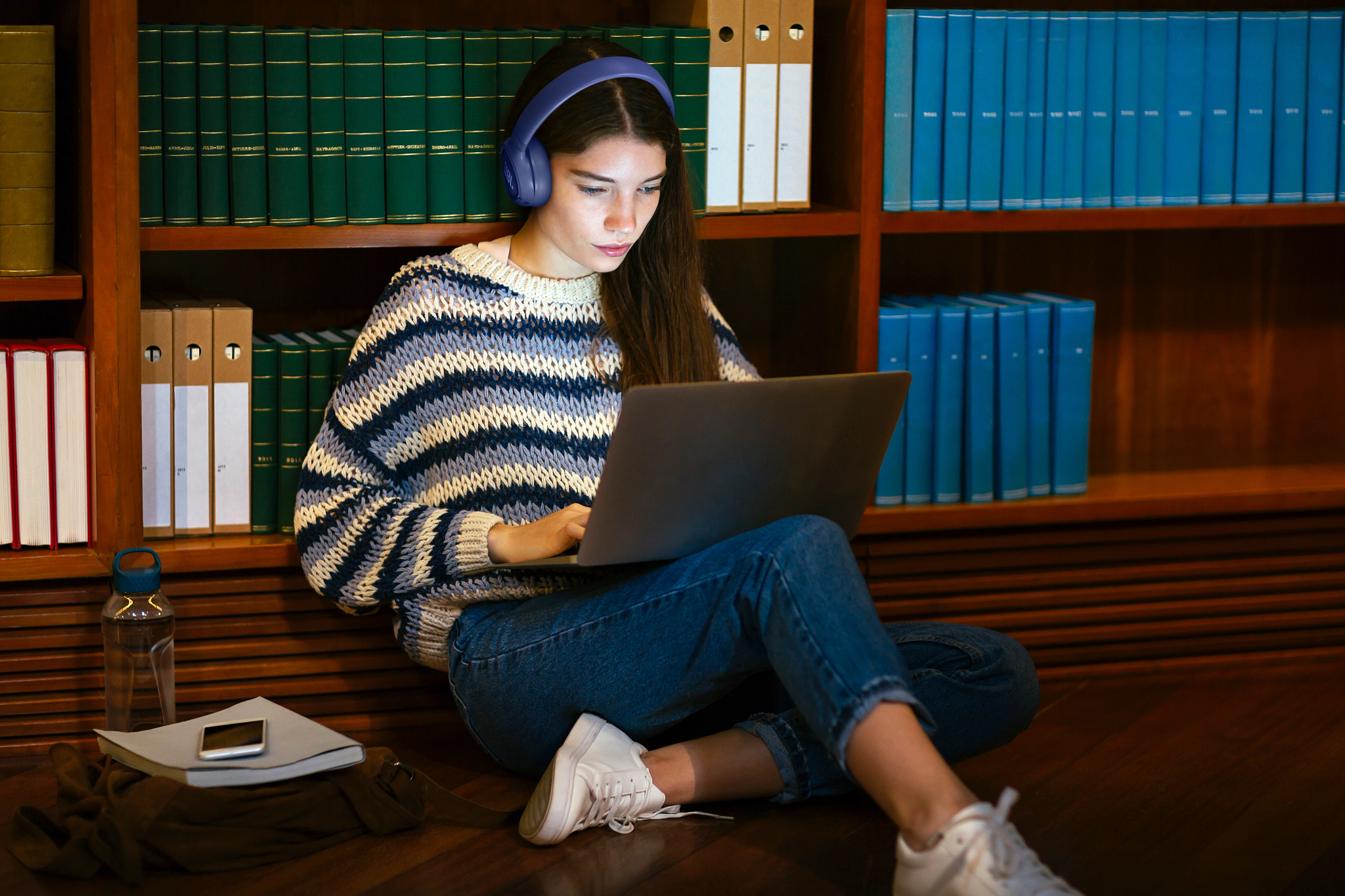 Junge Frau mit Kopfhörern lernt auf einem Laptop vor einem Bücherregal.
