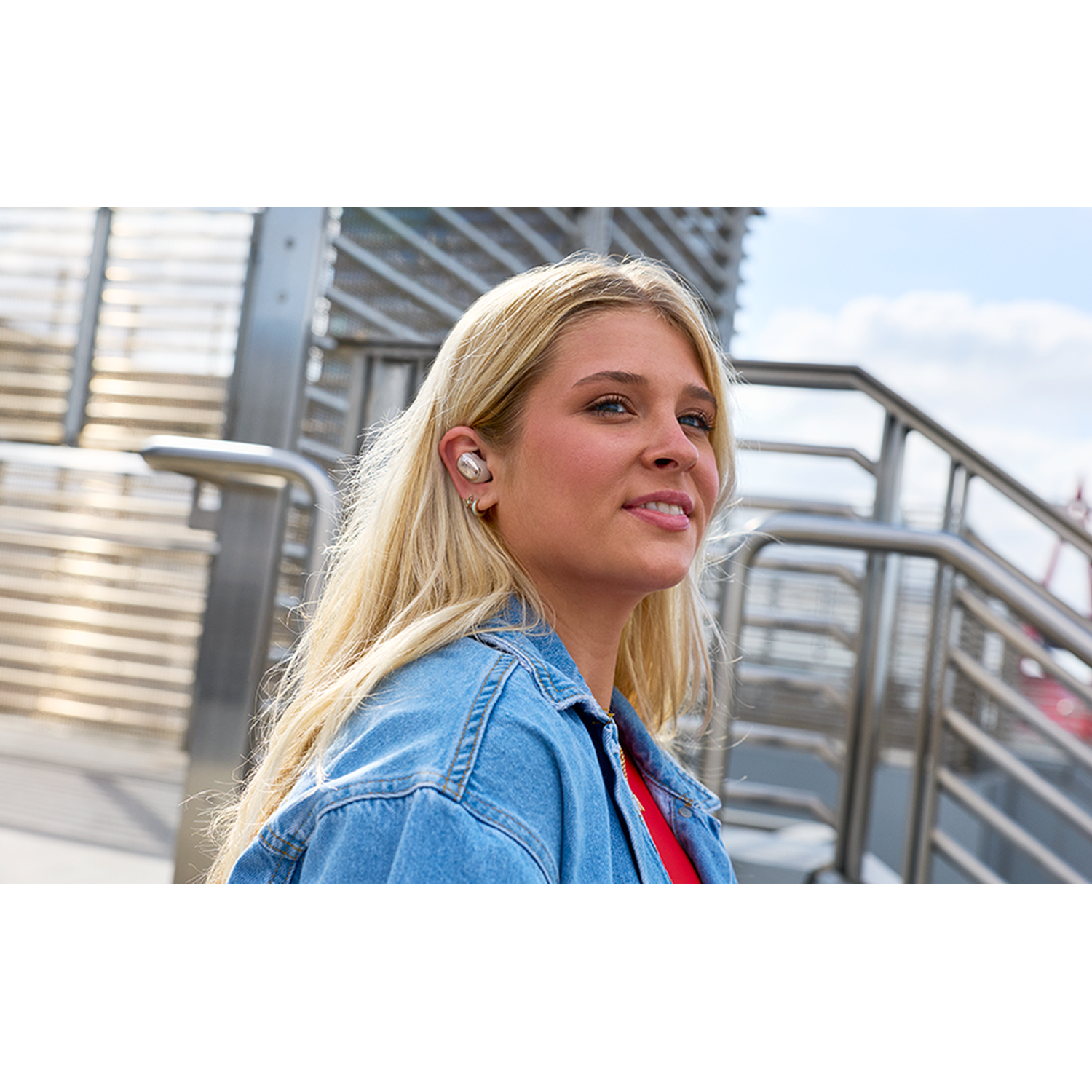 Blondine in spijkerjasje, met oordopjes, kijkt naar rechts. Metalen trap erachter.