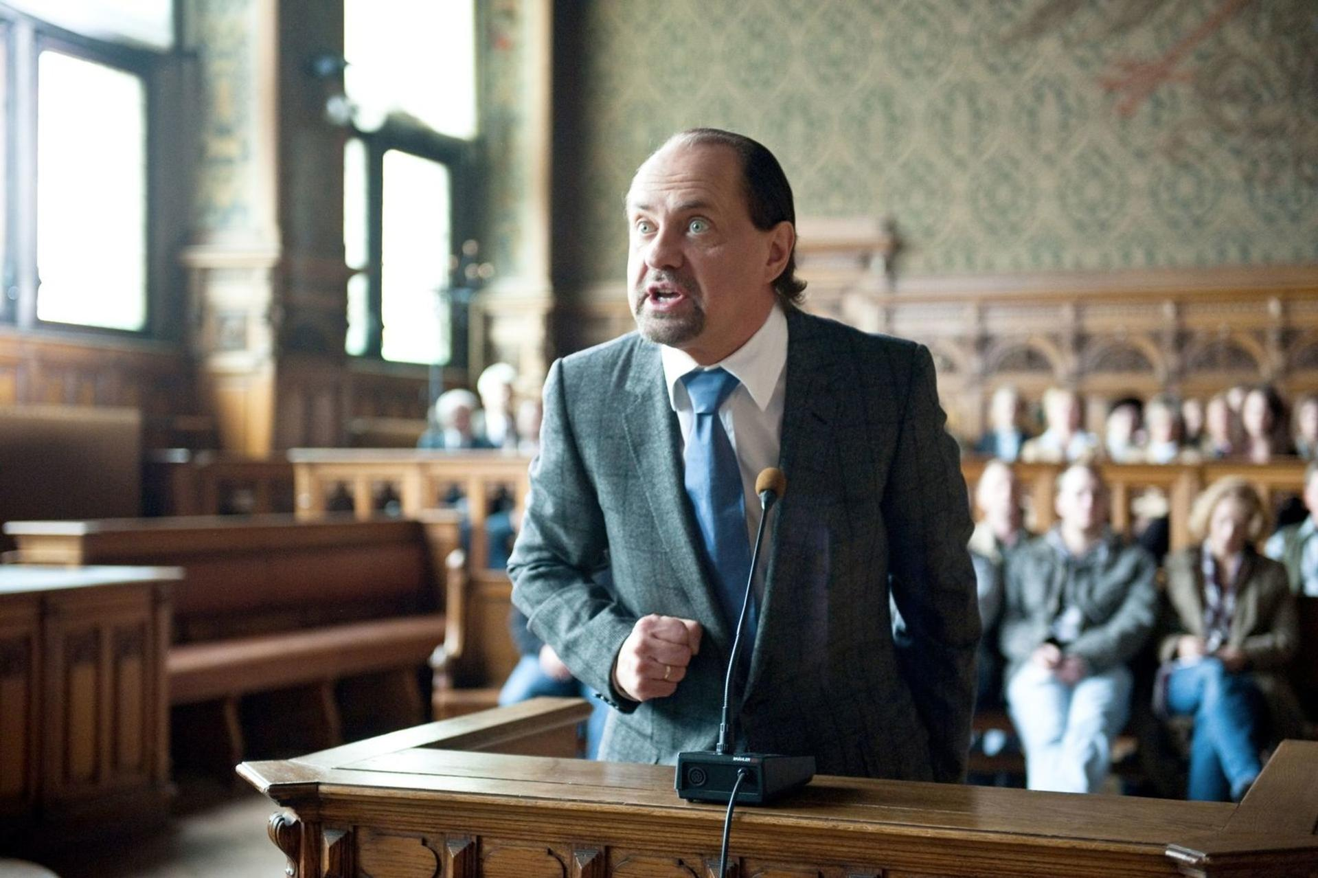 Ein Mann in einem Anzug spricht in ein Mikrofon, mit Zuhörern im Hintergrund.