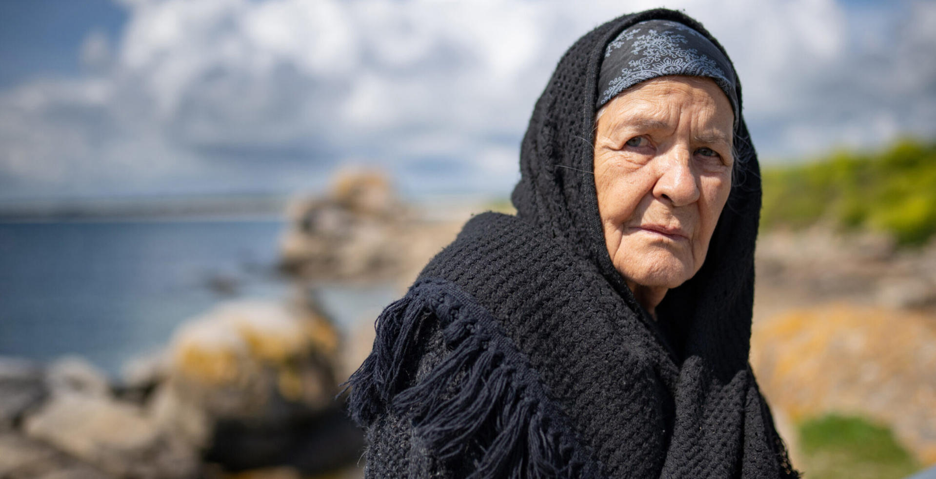 Ältere Frau in schwarzem Kopftuch und Strickjacke am Meer.