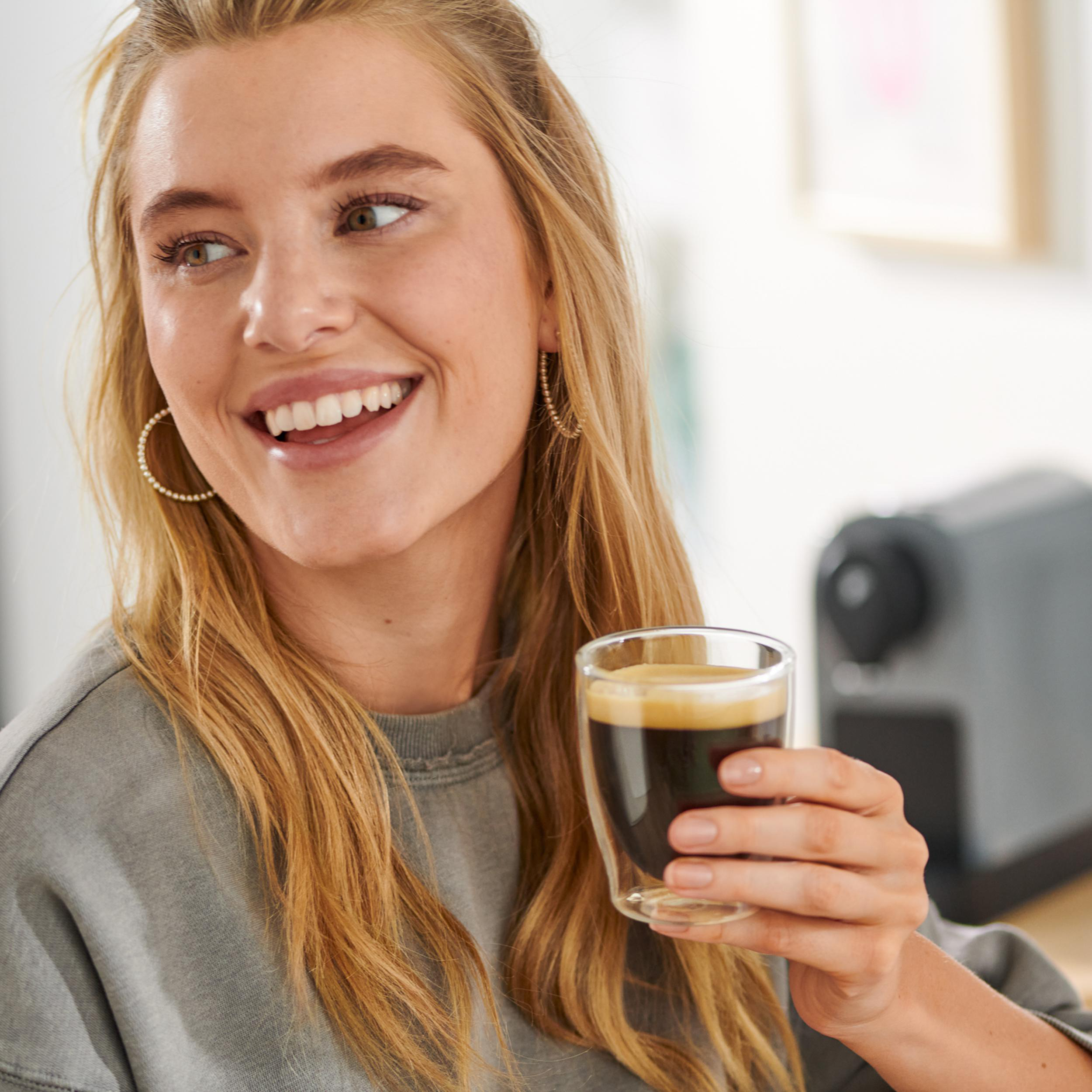 Eine Frau mit blonden Haaren hält ein Glas Espresso. Eine Kaffeemaschine ist im Hintergrund.
