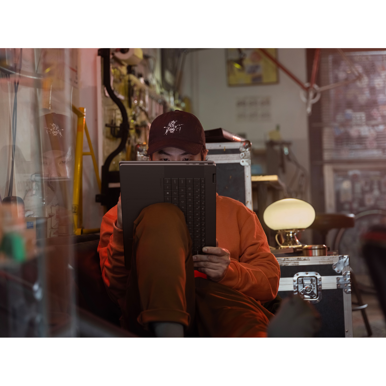 Man in cap and orange sweater typing on a laptop resting on his lap.