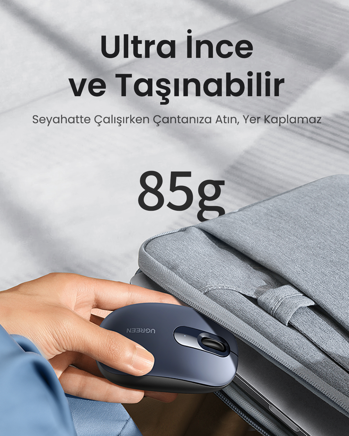 Bir el, gri bir dizüstü bilgisayar çantasının yanında koyu renkli bir fare tutuyor. Metin: 85g.