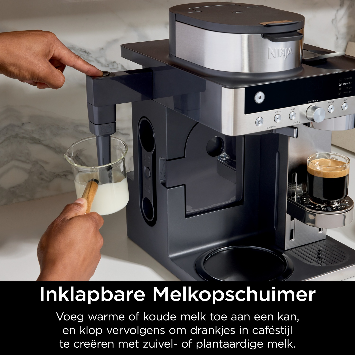 A hand presses a button on a coffee machine while milk is frothed in a pitcher.