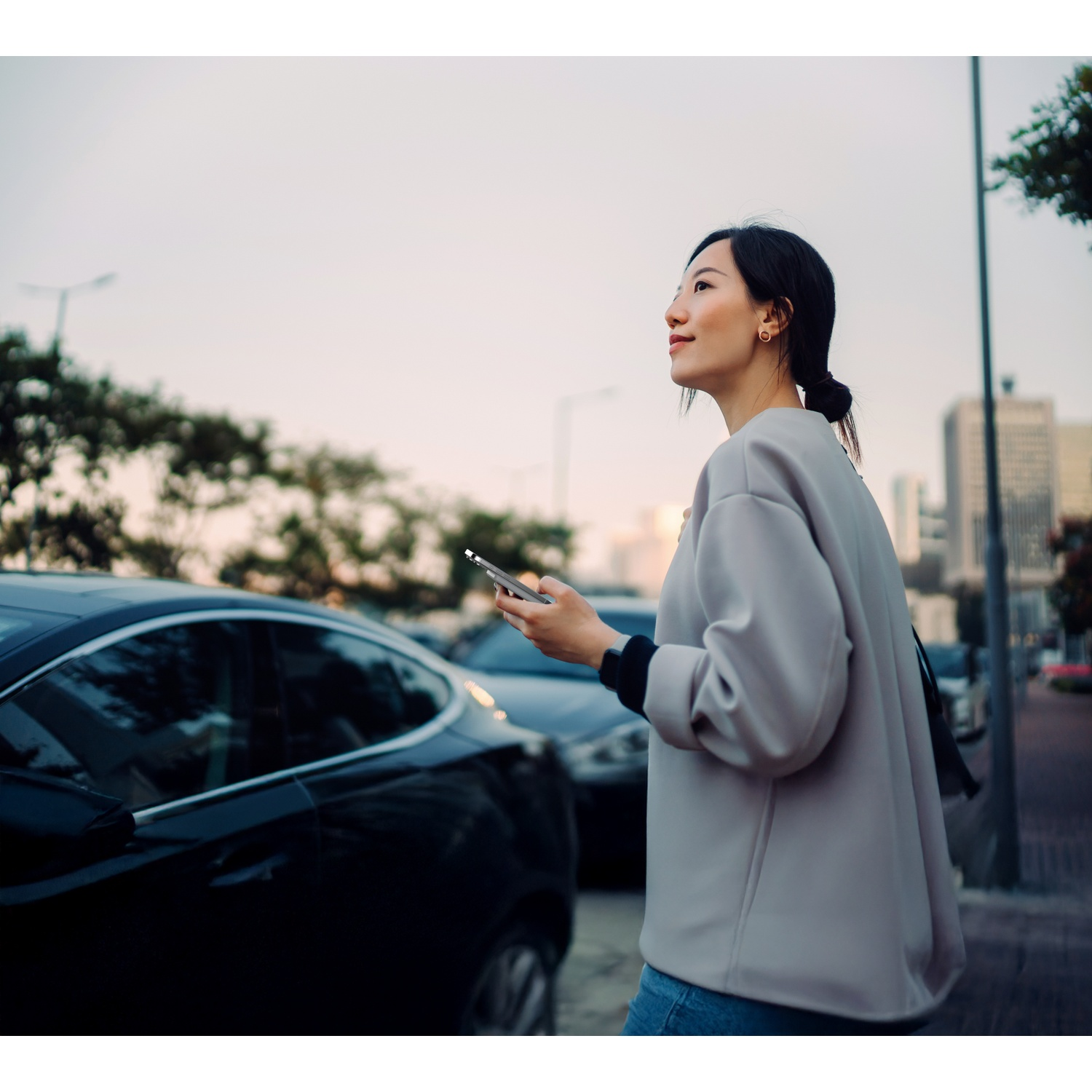 Femme tenant un téléphone près de voitures garées dans une rue de ville au crépuscule.