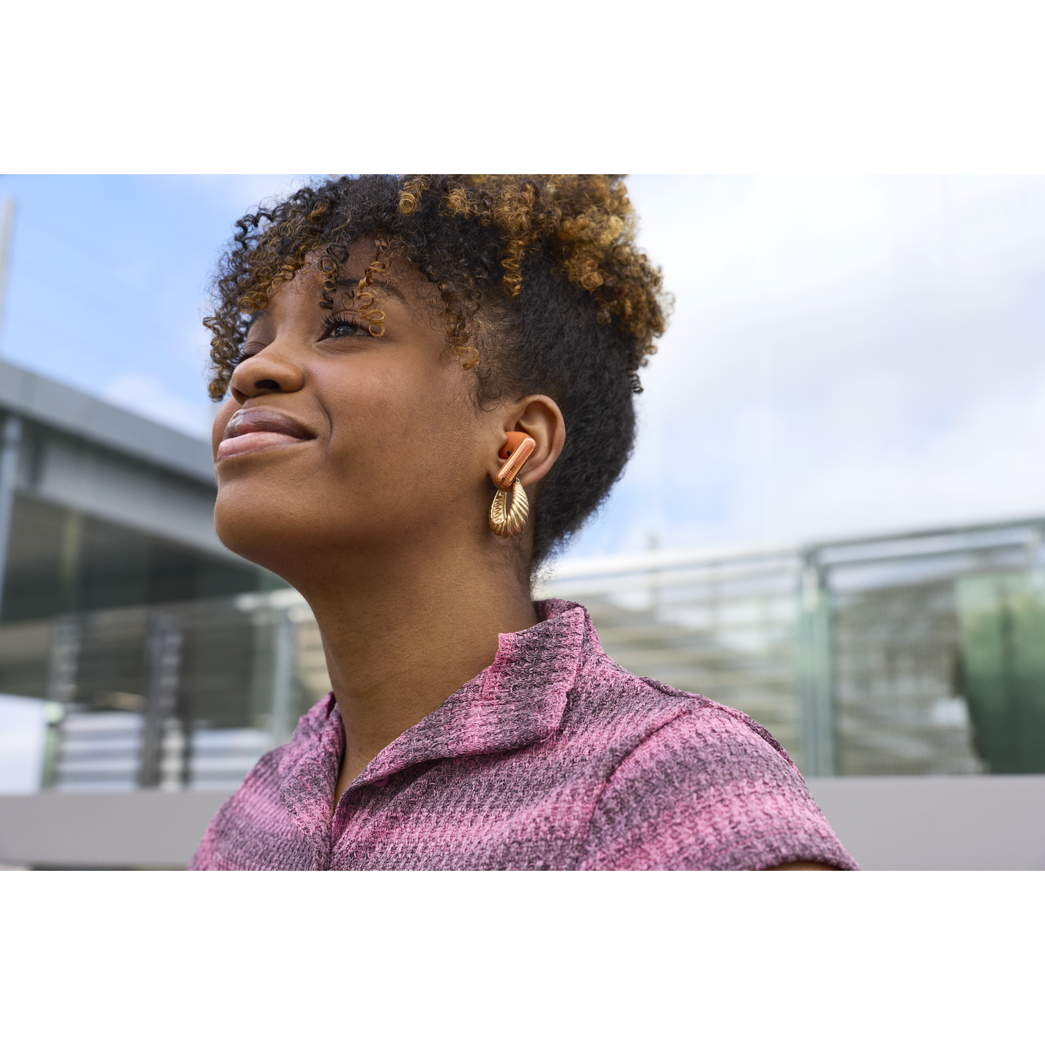 Femme aux cheveux bouclés et écouteurs orange, porte une chemise rose, regarde en l'air.
