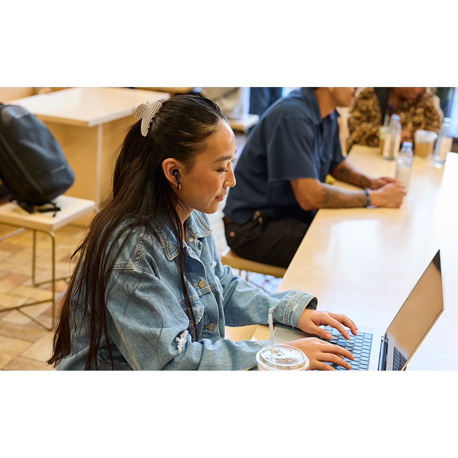A young woman in a denim jacket types on a laptop at a table with others.