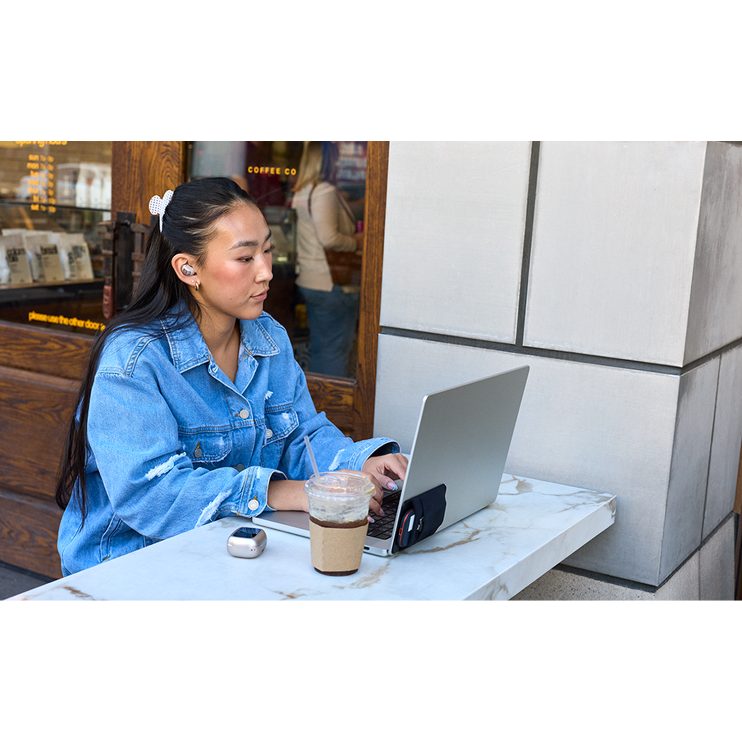 Une jeune femme en veste en jean travaille sur un ordinateur portable, assise dehors à une table avec un café.