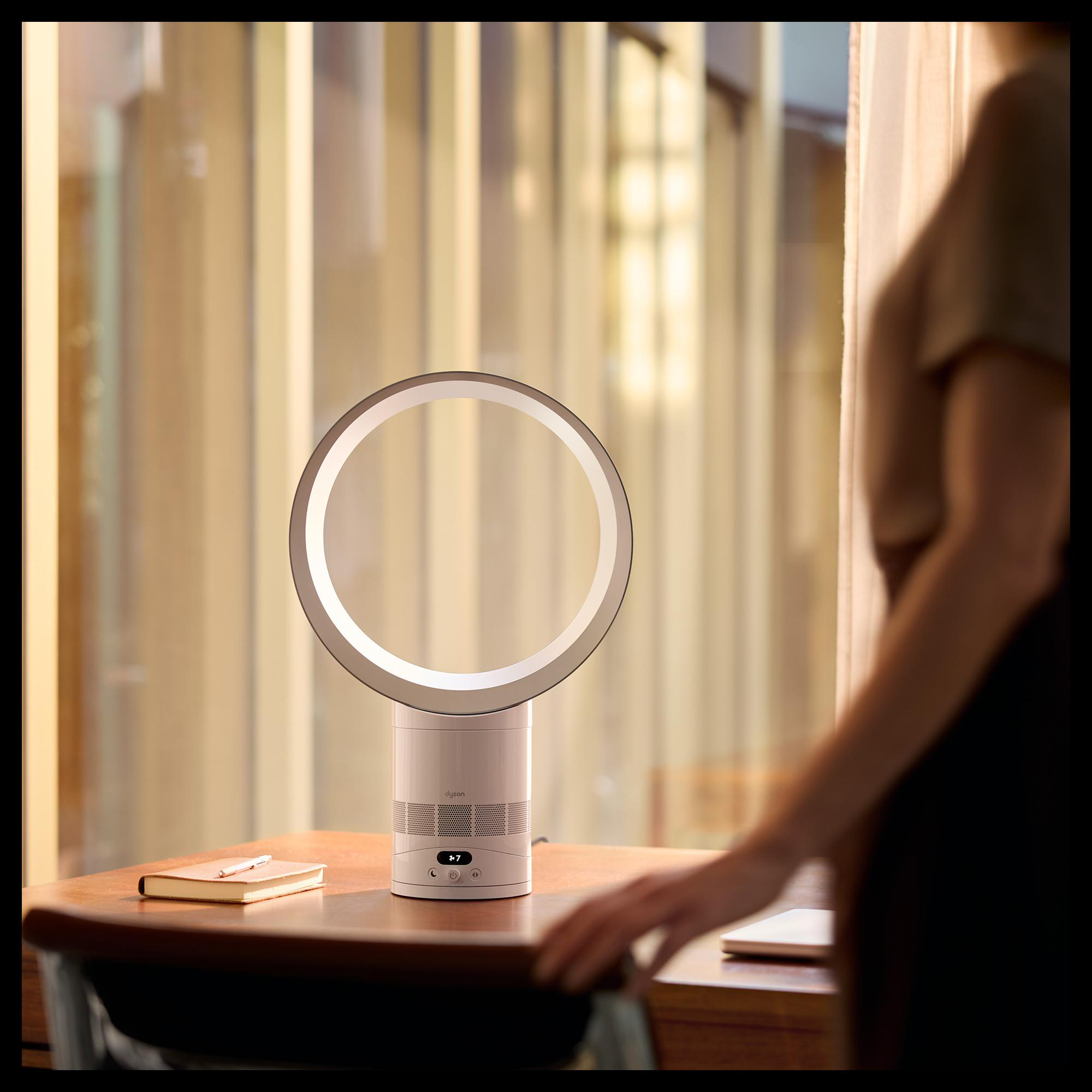 Ein runder, weißer Ventilator steht auf einem Holztisch. Eine Frau ist rechts unscharf.