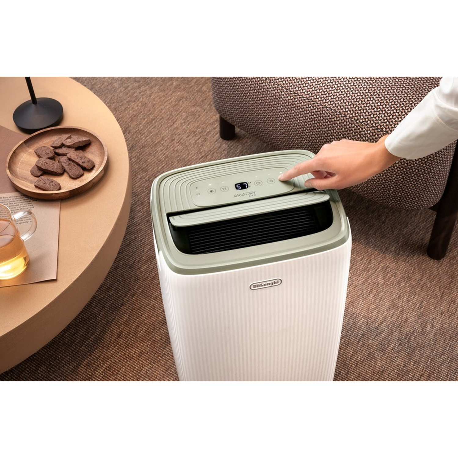 Una mano preme un pulsante su un deumidificatore bianco DeLonghi accanto a un tavolo con biscotti e tè.