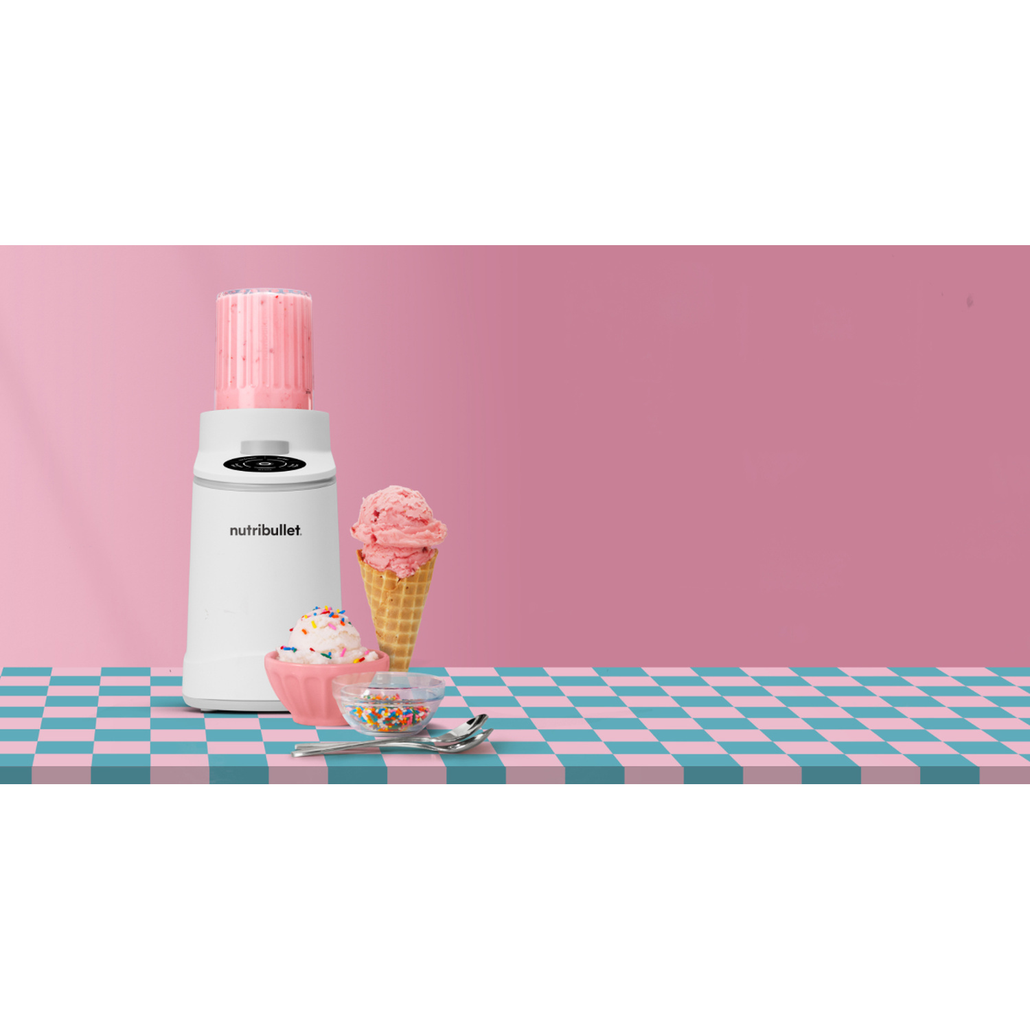 Un frullatore bianco con frullato rosa, gelato e zuccherini su un tavolo a scacchi.