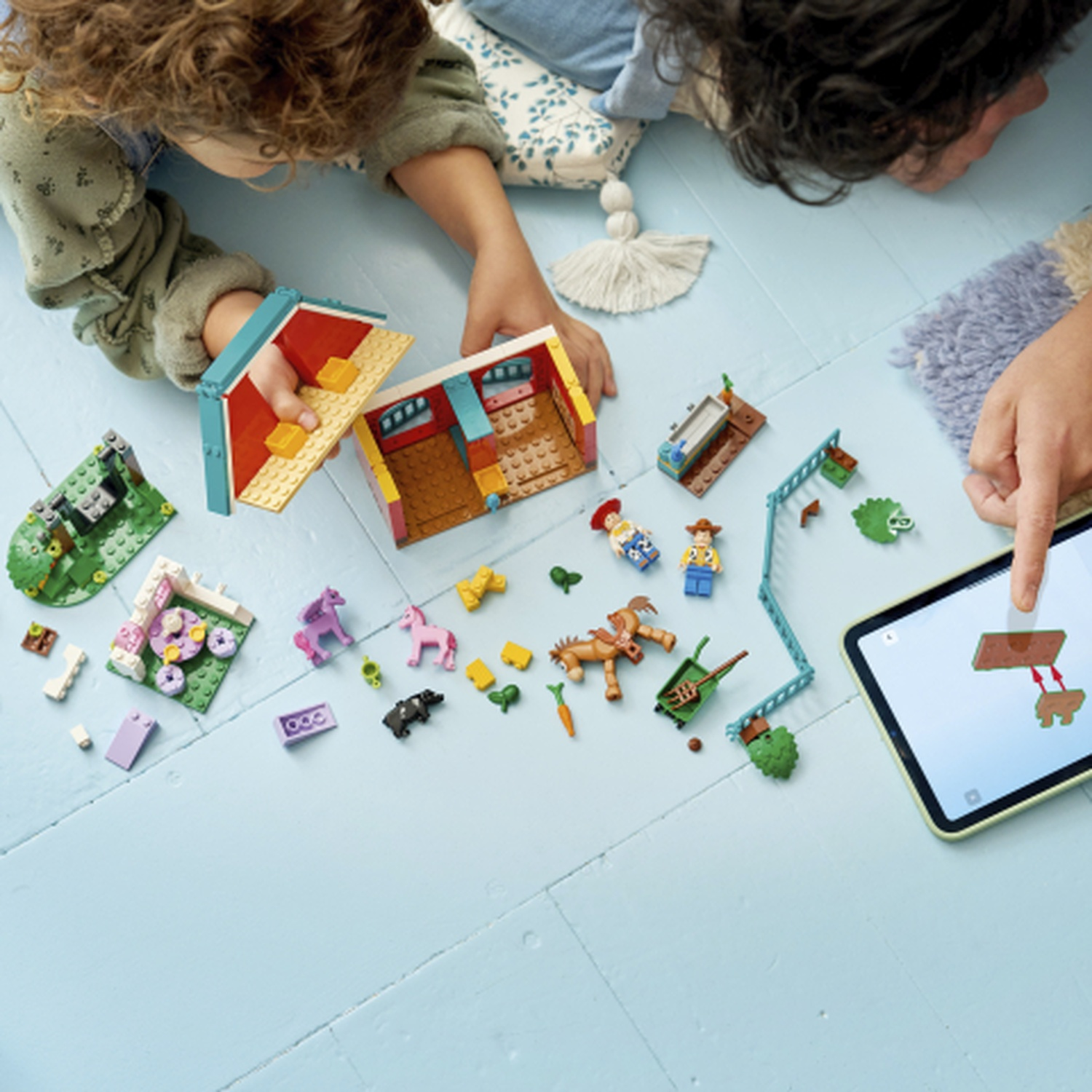 Bambini giocano con giocattoli di fattoria Lego, costruendo una casa e animali da fattoria. Un tablet mostra istruzioni.