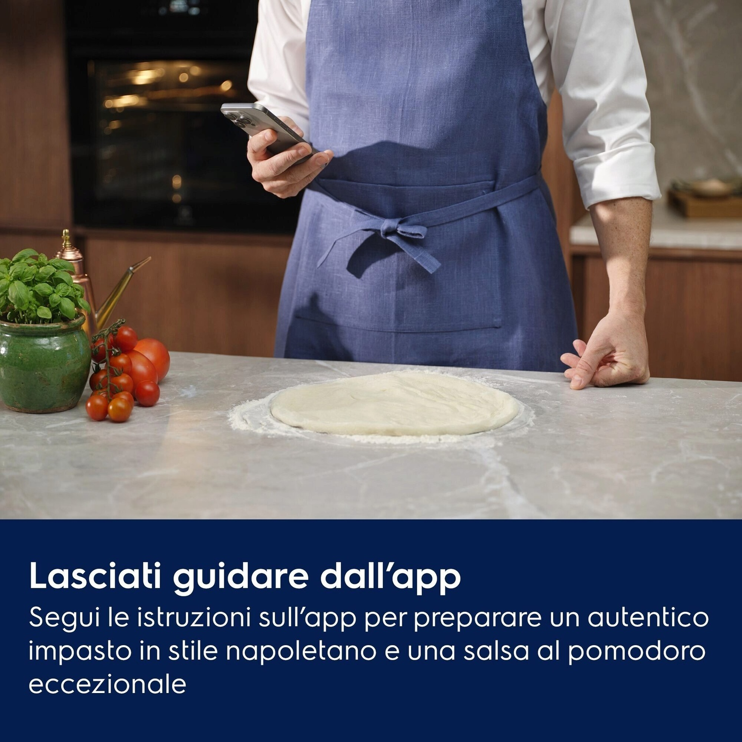 Uomo con smartphone in cucina prepara impasto, pomodori e basilico vicini.