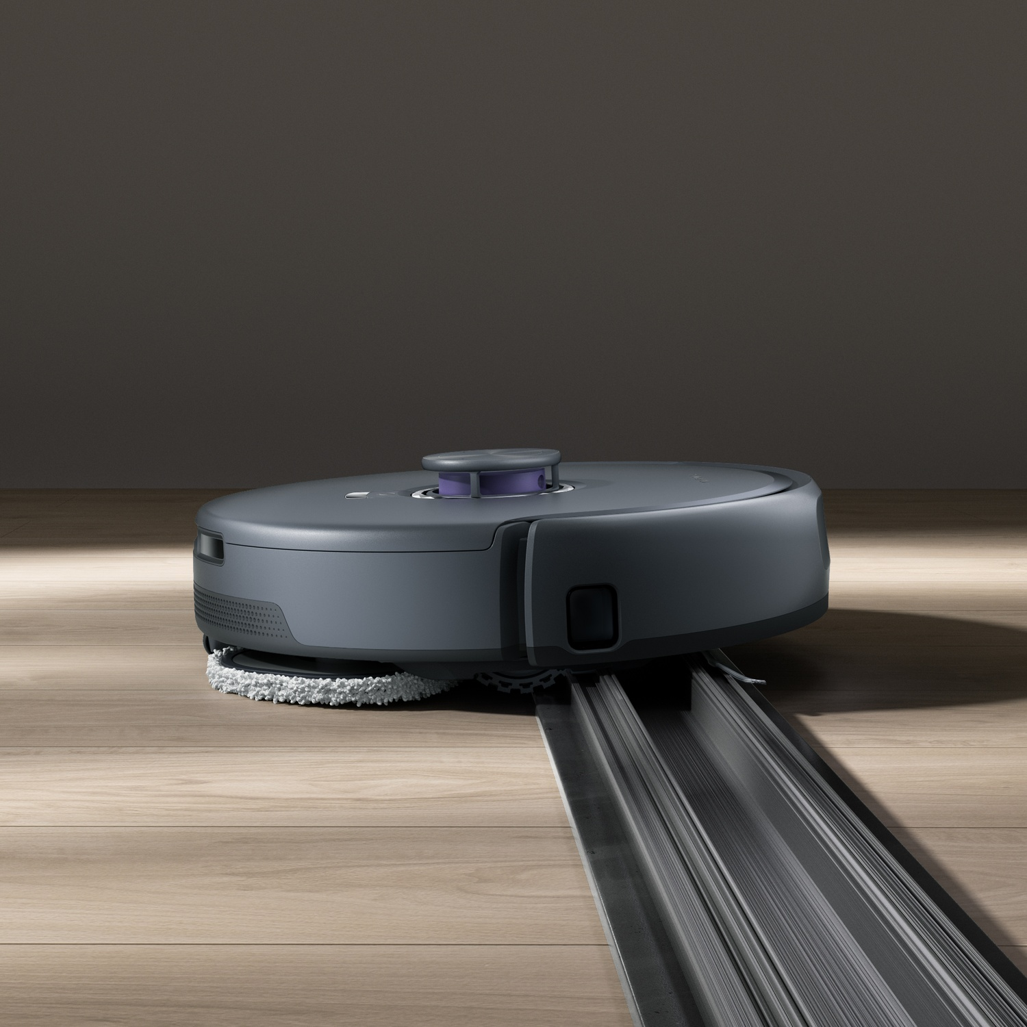 Un robot aspirapolvere grigio con funzione lavaggio si muove su un pavimento in legno sopra un binario metallico.