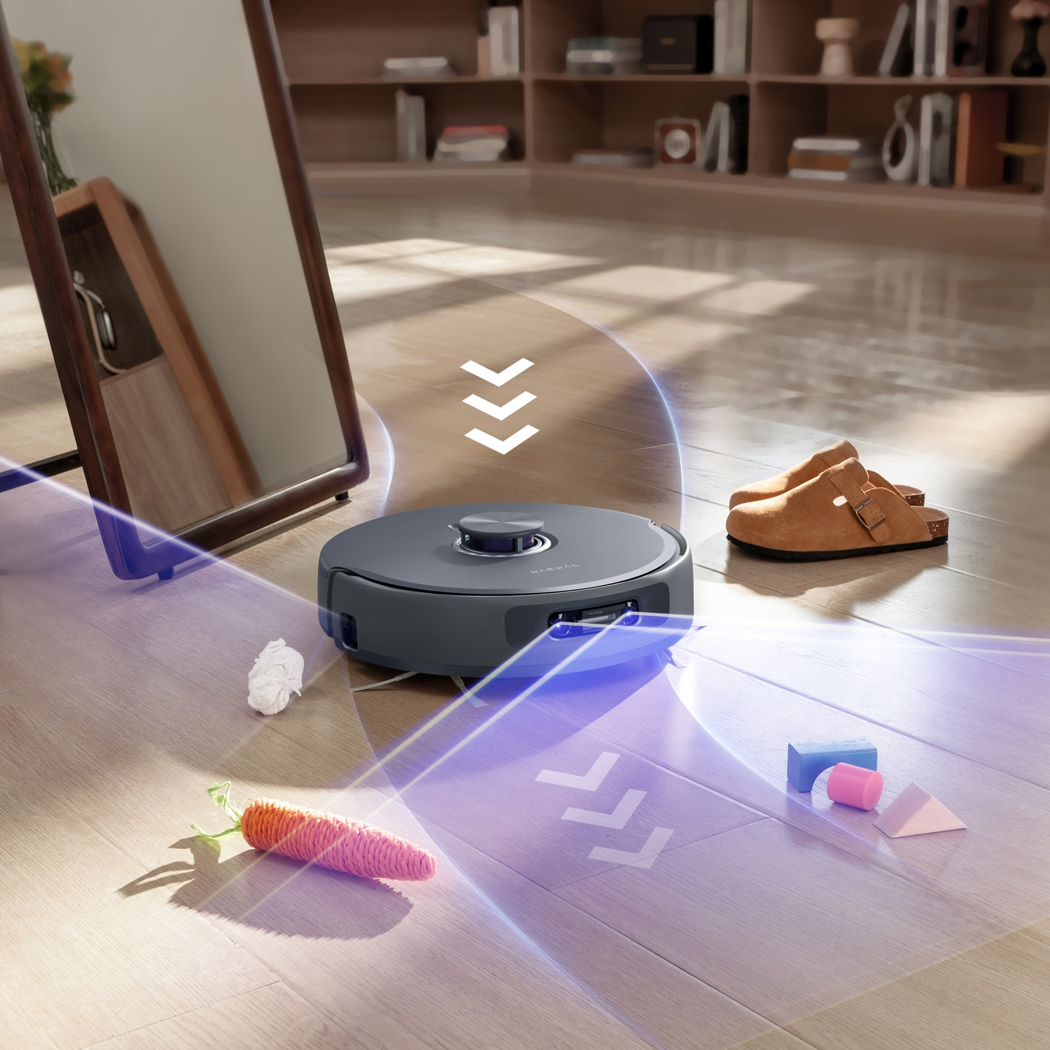 Un robot aspirapolvere pulisce un pavimento in legno vicino a uno specchio, pantofole e giocattoli.