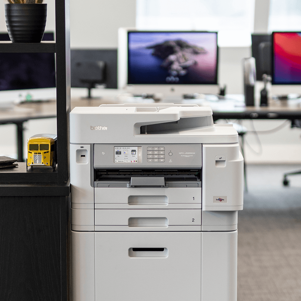 Biała drukarka Brother stoi na biurku w biurze z komputerami w tle.
