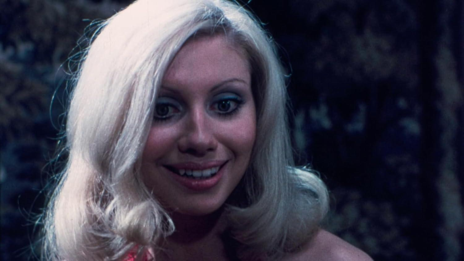 Eine blonde Frau mit blauen Lidschatten lächelt in die Kamera. Dunkler Hintergrund.