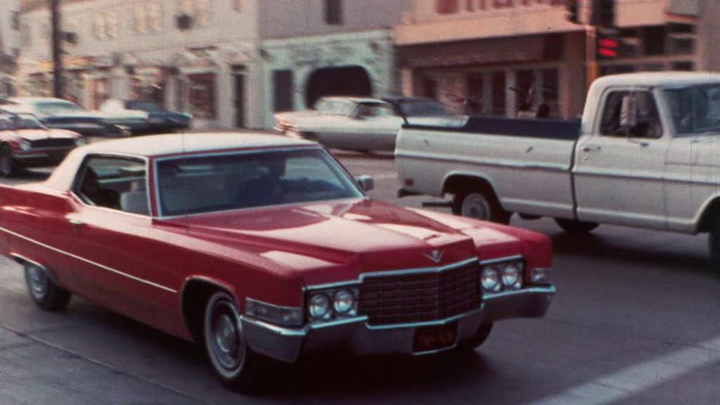 Ein roter Cadillac fährt auf einer Straße mit anderen Autos und Gebäuden im Hintergrund.