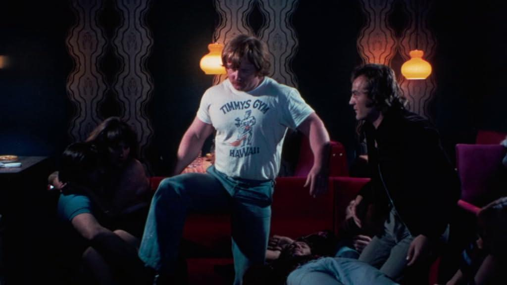 Ein Mann mit einem "Timmy's Gym Hawaii" T-Shirt steht. Mehrere Personen liegen auf Sofas.