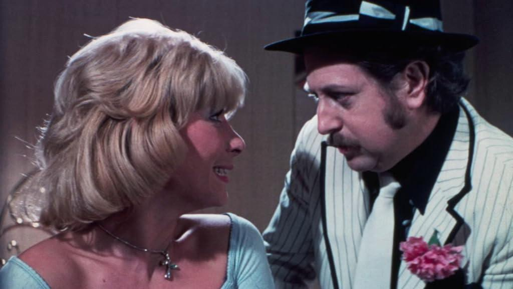 Eine blonde Frau und ein Mann im Nadelstreifenanzug und Hut sprechen.