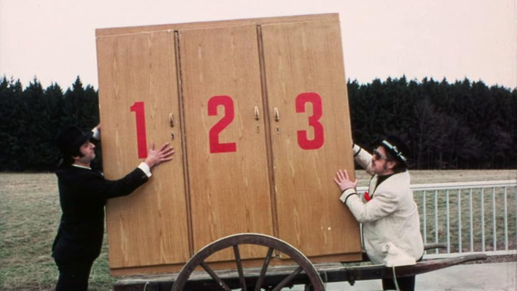 Zwei Männer schieben einen großen Holzschrank mit den Zahlen 1, 2, 3 darauf.