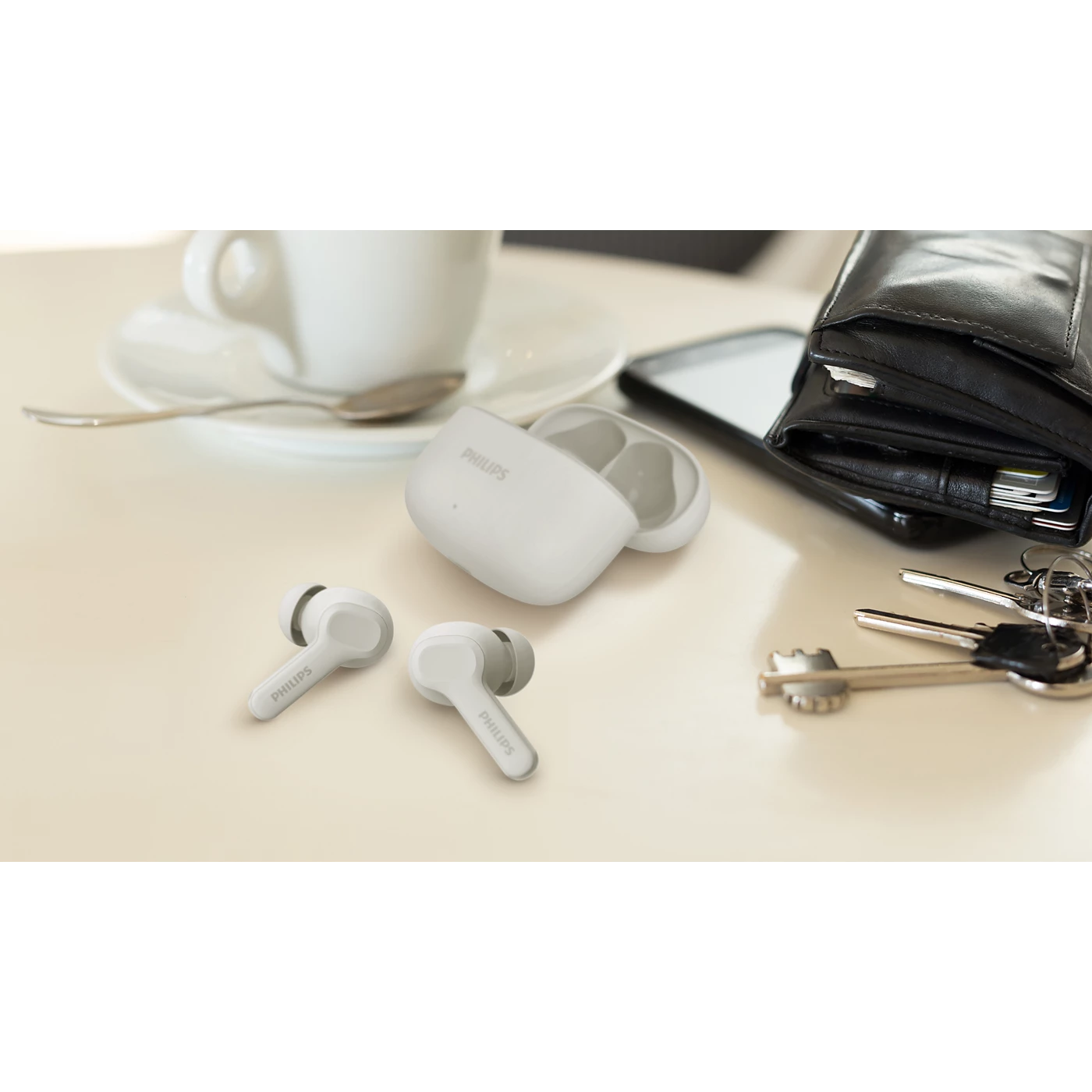 Beyaz Philips kulaklık ve kılıf, kahve fincanı, cüzdan, telefon ve anahtarlarla masada.