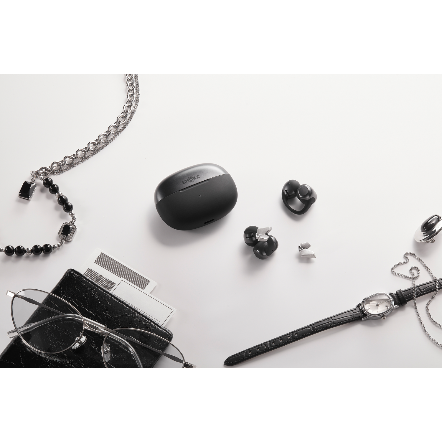 Schwarze kabellose Ohrhörer, Brille, Uhr und Schmuck auf weißem Untergrund.