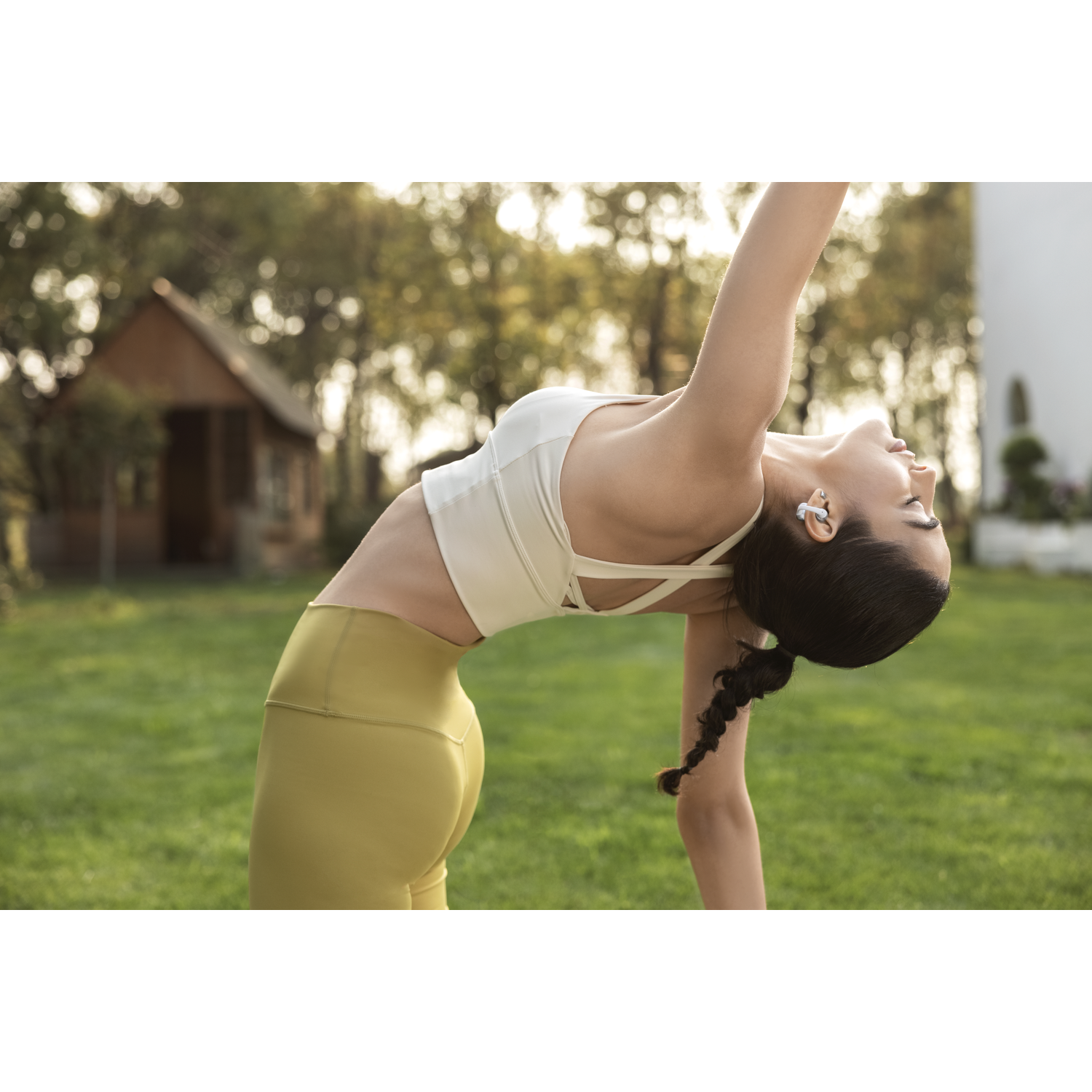 Frau in weißem Oberteil und gelber Leggings macht Yoga im Gras mit kleinem Haus.