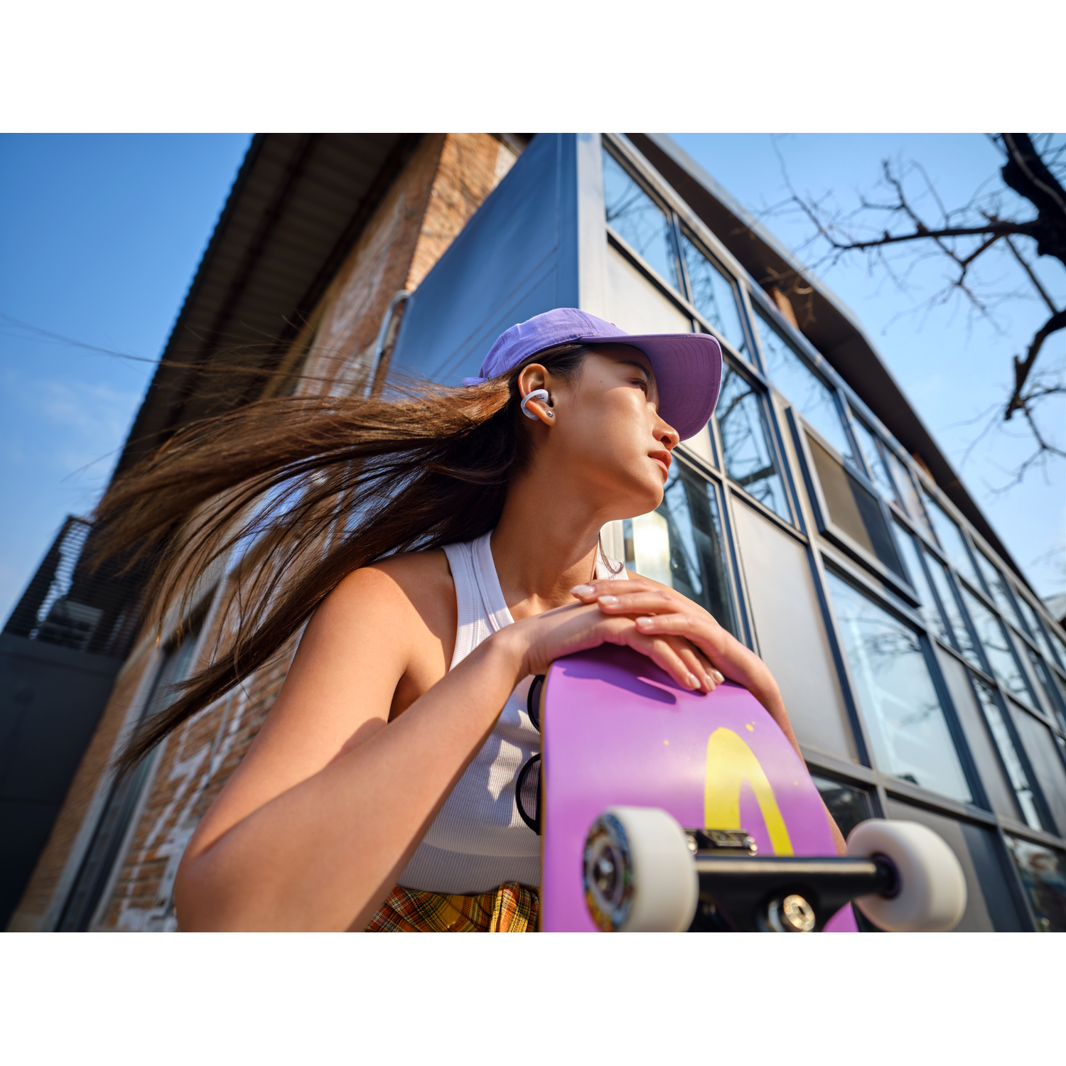 Young woman with purple cap and white top holds purple skateboard. Building background.