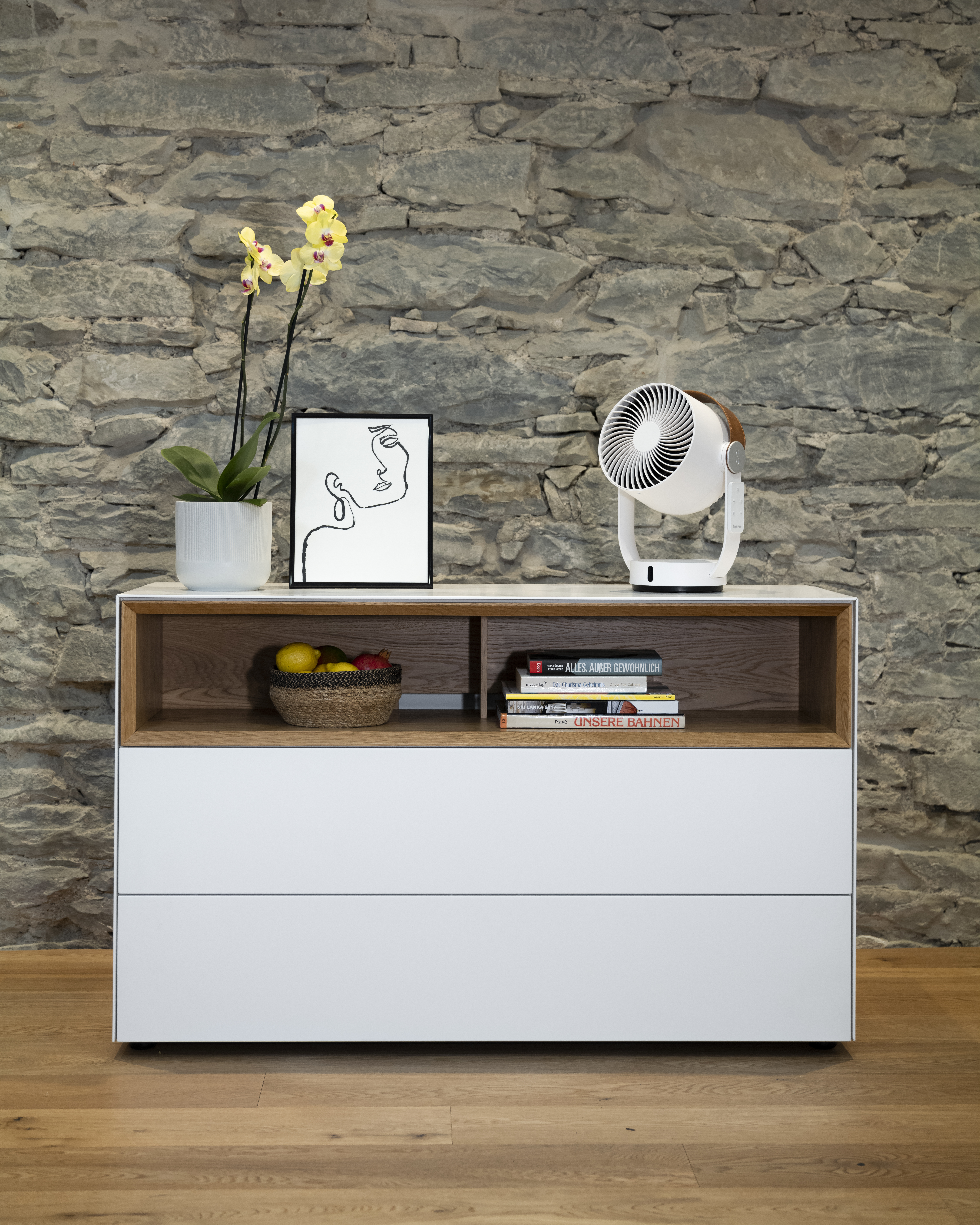 Eine weiße Kommode mit einer Orchidee, einem Kunstwerk und einem Ventilator auf dem Tisch.