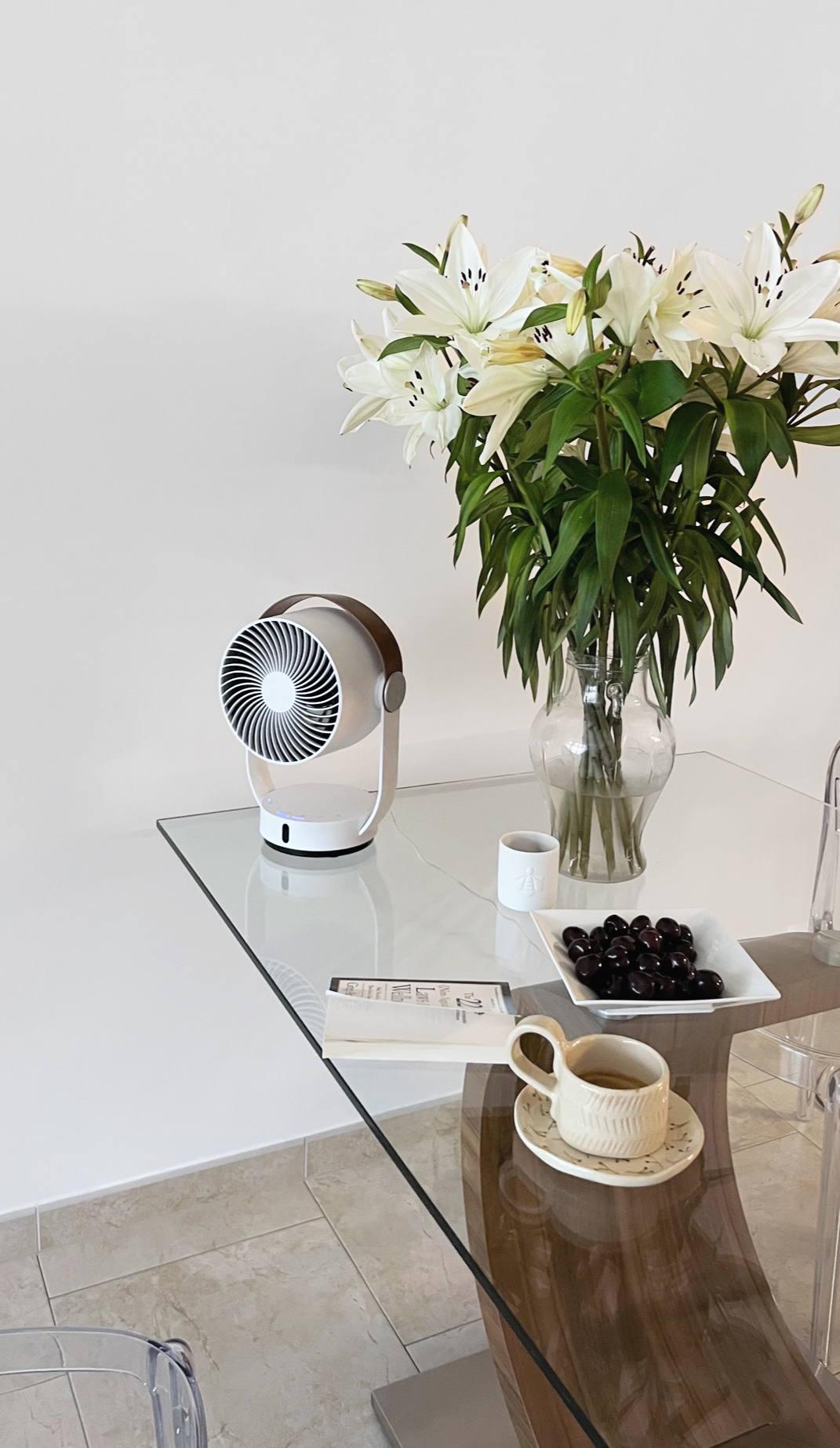 Ein kleiner Ventilator, eine Vase mit weißen Lilien, eine Schale mit Kirschen und eine Tasse Kaffee auf einem Glastisch.
