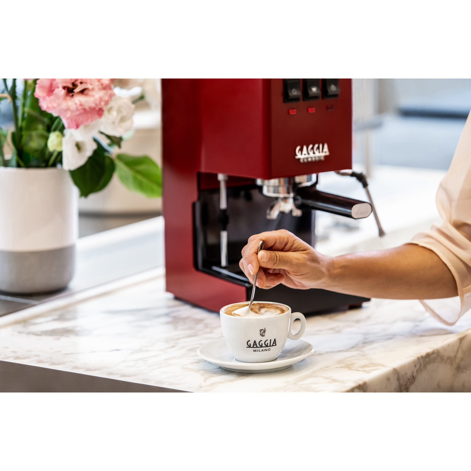 Una persona mescola un caffè in una tazza bianca accanto a una macchina per espresso rossa.
