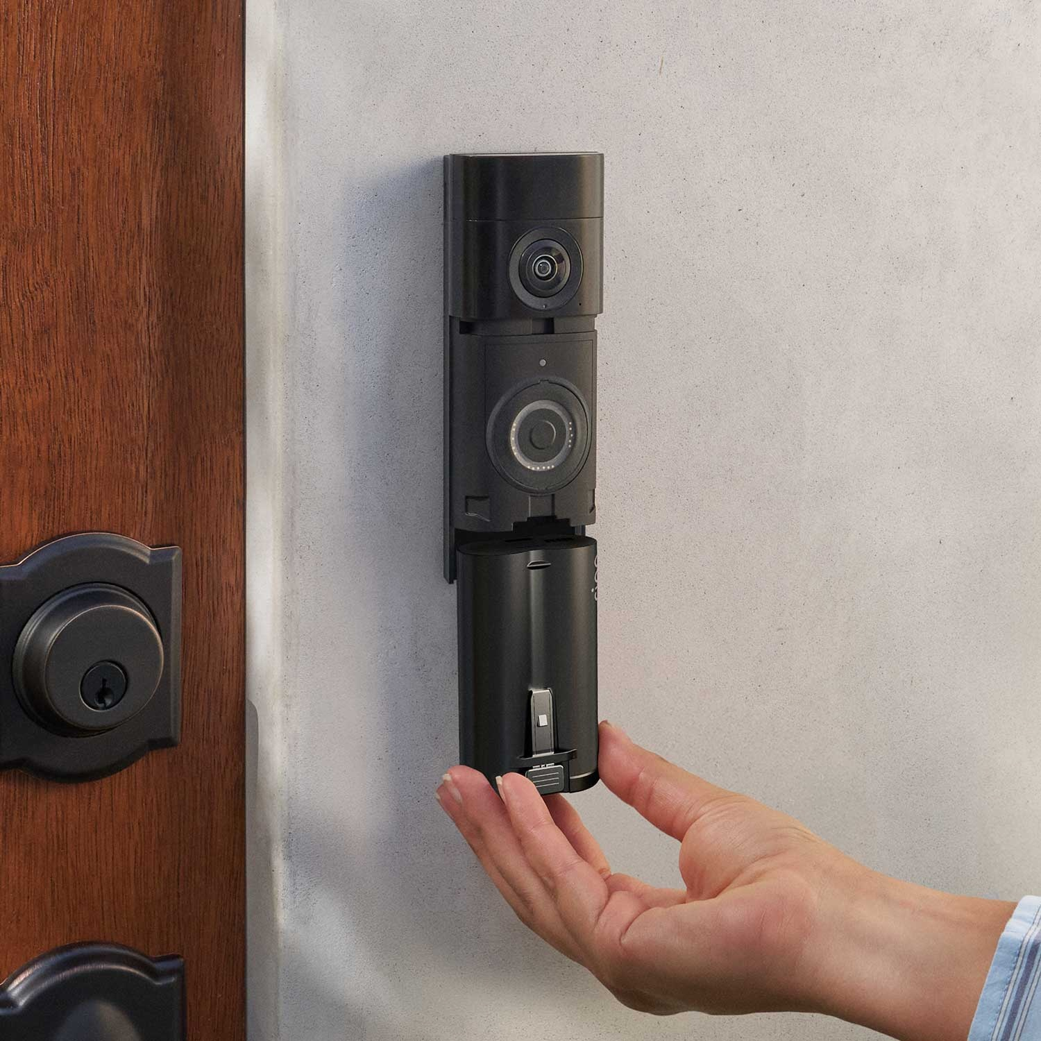 Une main place une caméra de sonnette noire sur un mur à côté d'une porte en bois.