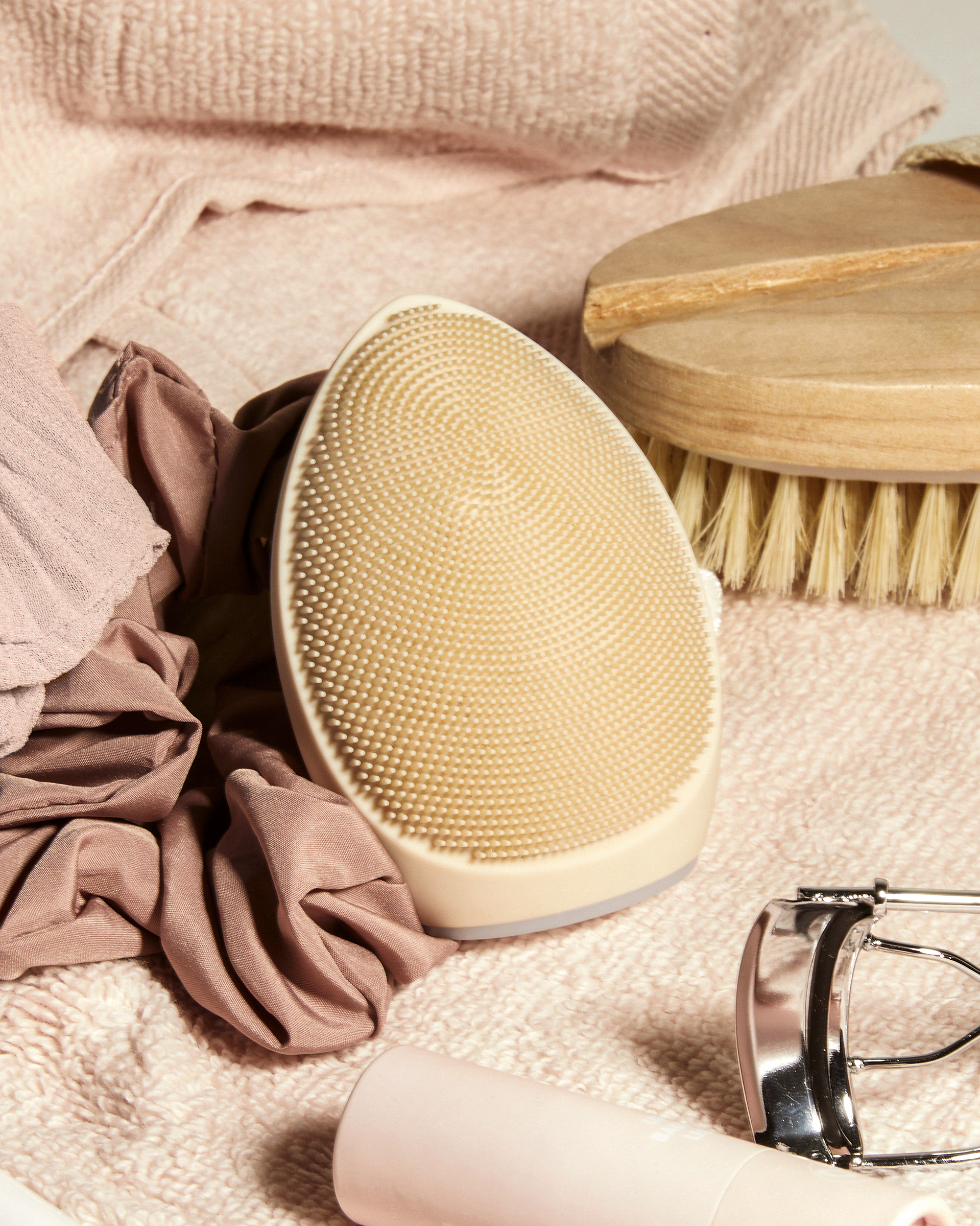 Un limpiador facial de silicona rosa claro junto a un cepillo de madera sobre textiles rosas.