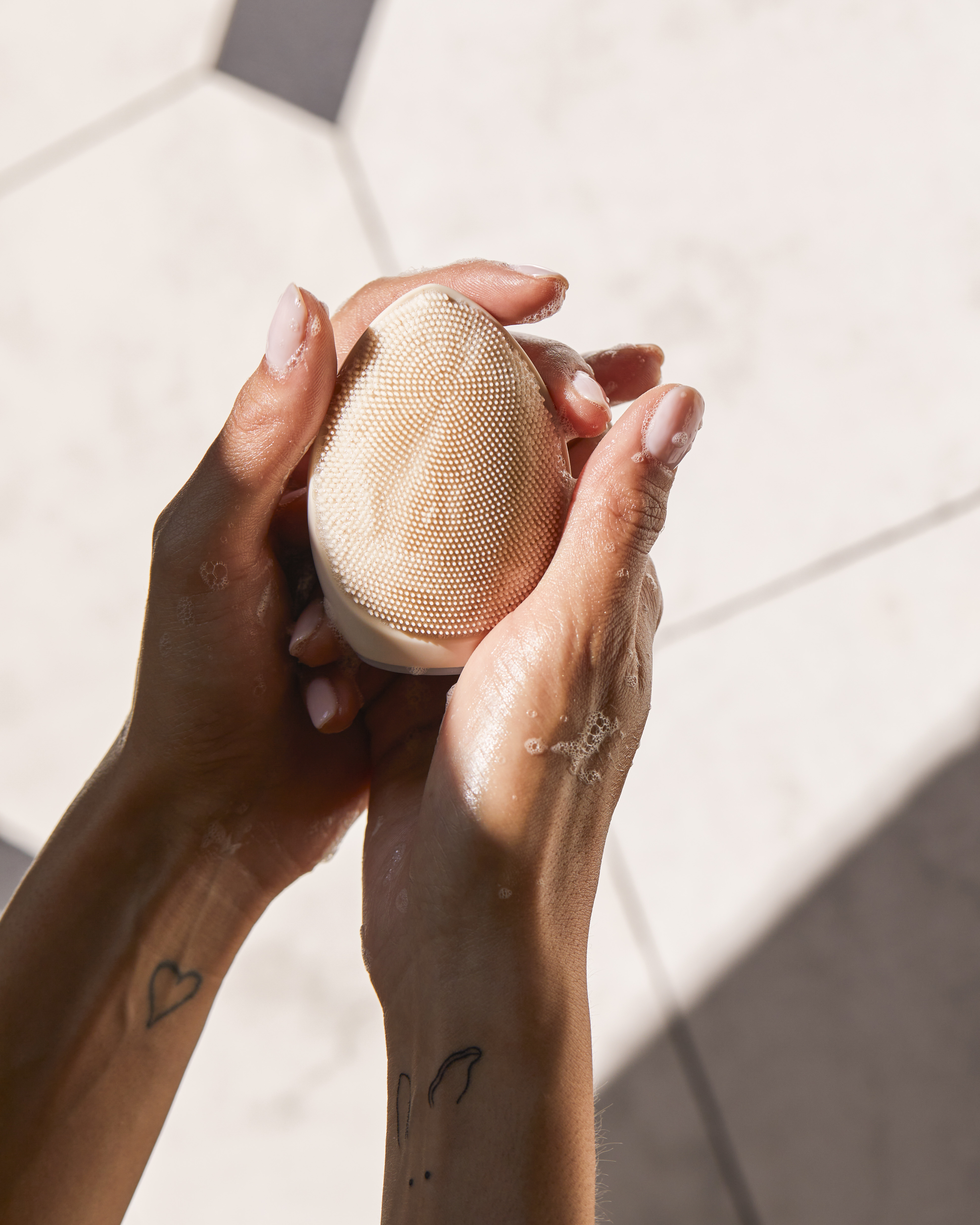 Manos sostienen un cepillo de limpieza facial beige con espuma de jabón.