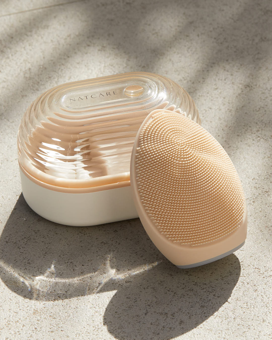 Un cepillo limpiador facial de silicona beige junto a su estuche con tapa transparente.