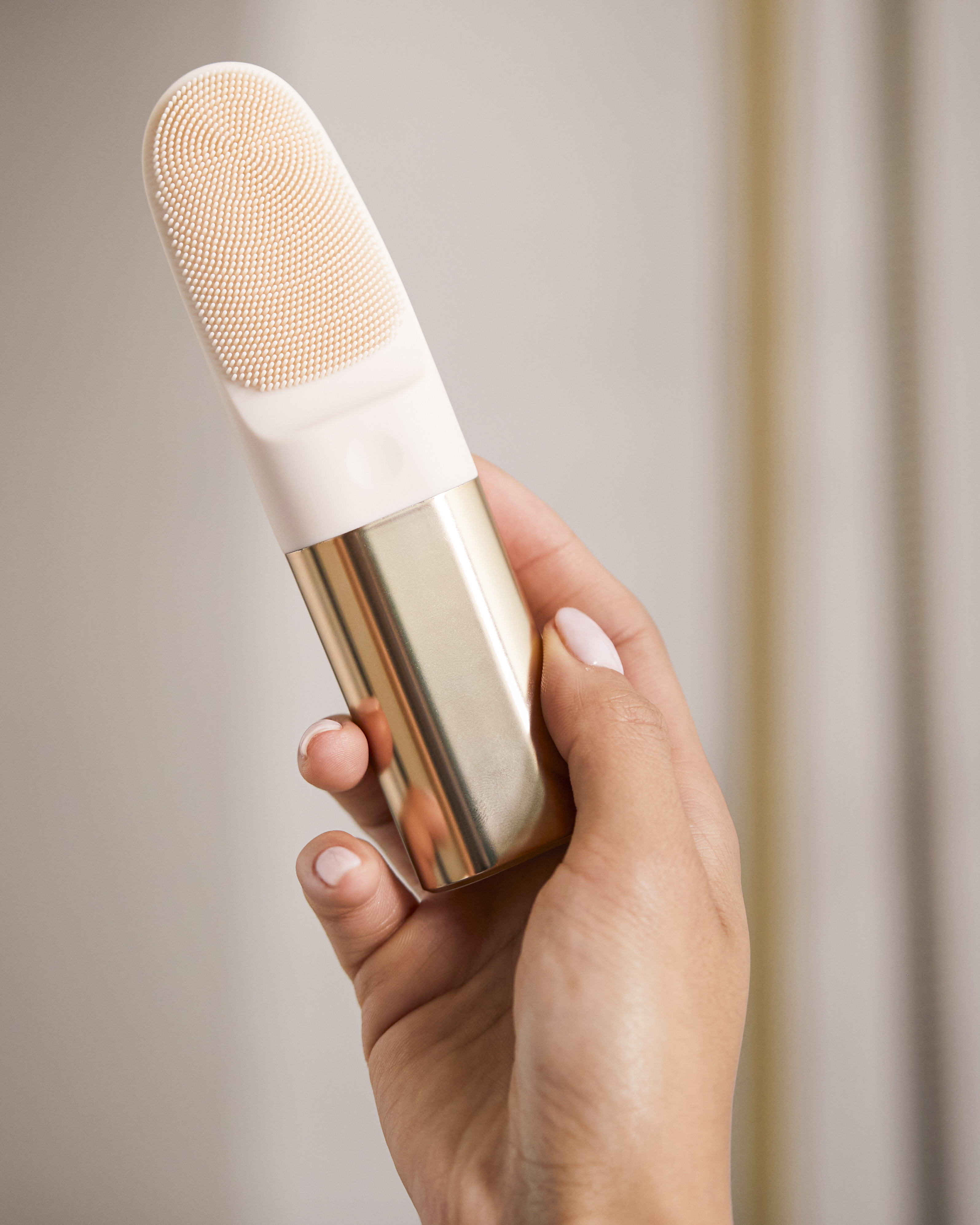 Una mano sostiene una herramienta de limpieza facial de silicona rosa con mango dorado.