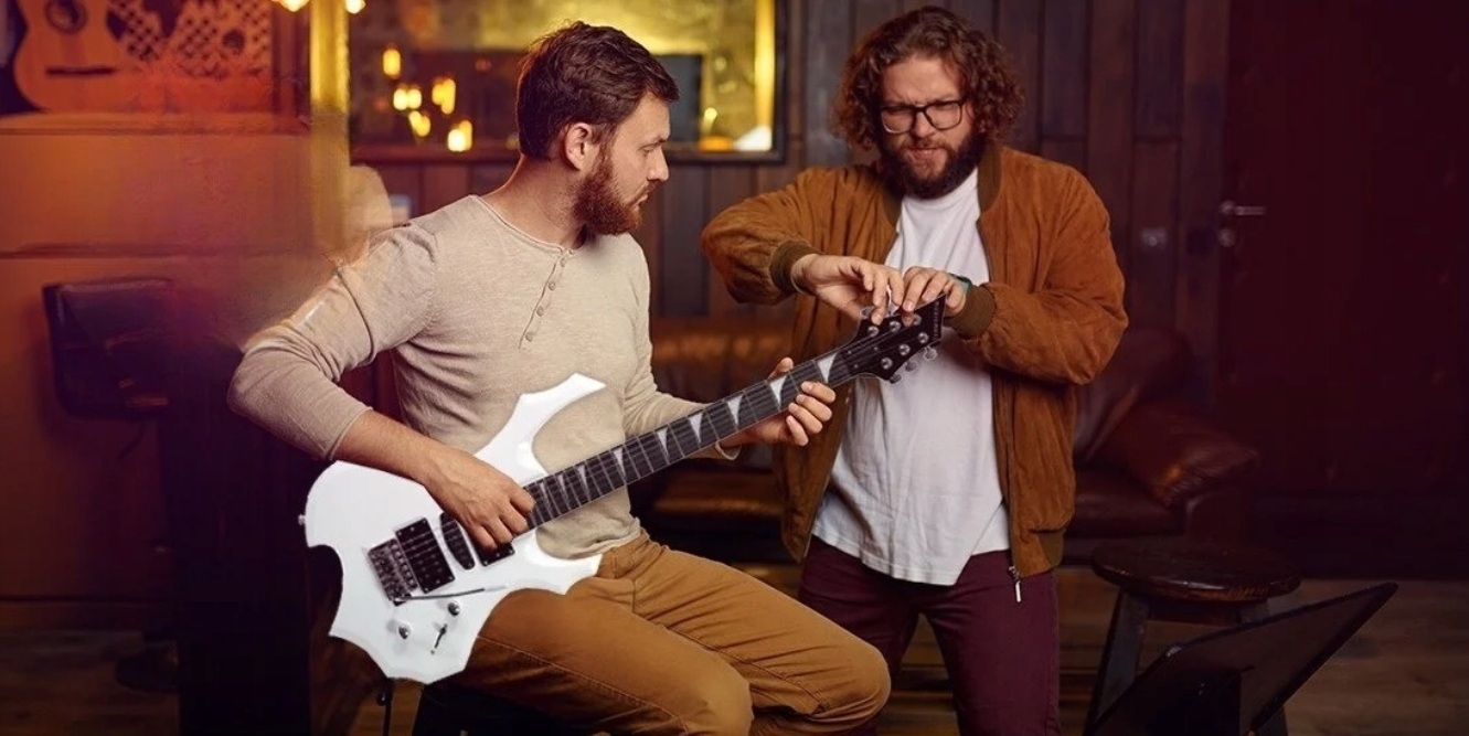 Mężczyzna stroi białą gitarę elektryczną, podczas gdy drugi mężczyzna obserwuje w słabo oświetlonym pomieszczeniu.