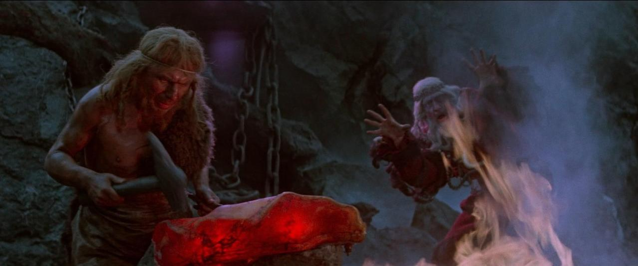 Ein Mann mit Fellkleidung bearbeitet glühendes Metall, ein anderer Mann mit Bart gestikuliert.