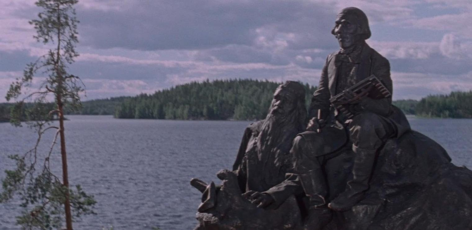 Skulptur von Männern an einem See mit Wald und bewölktem Himmel.