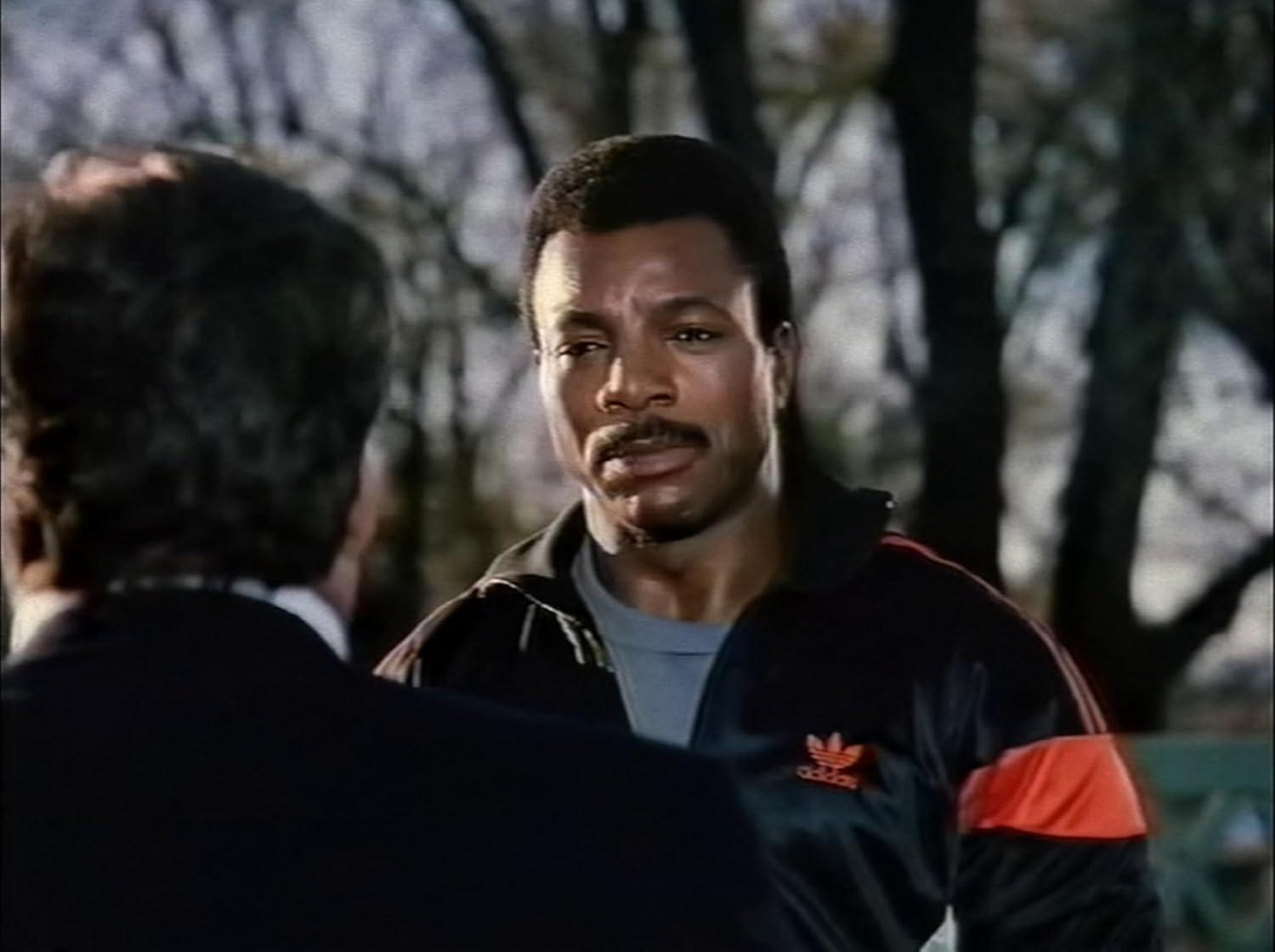 Ein Mann mit Schnurrbart in einem schwarzen Adidas Trainingsanzug spricht mit jemandem.