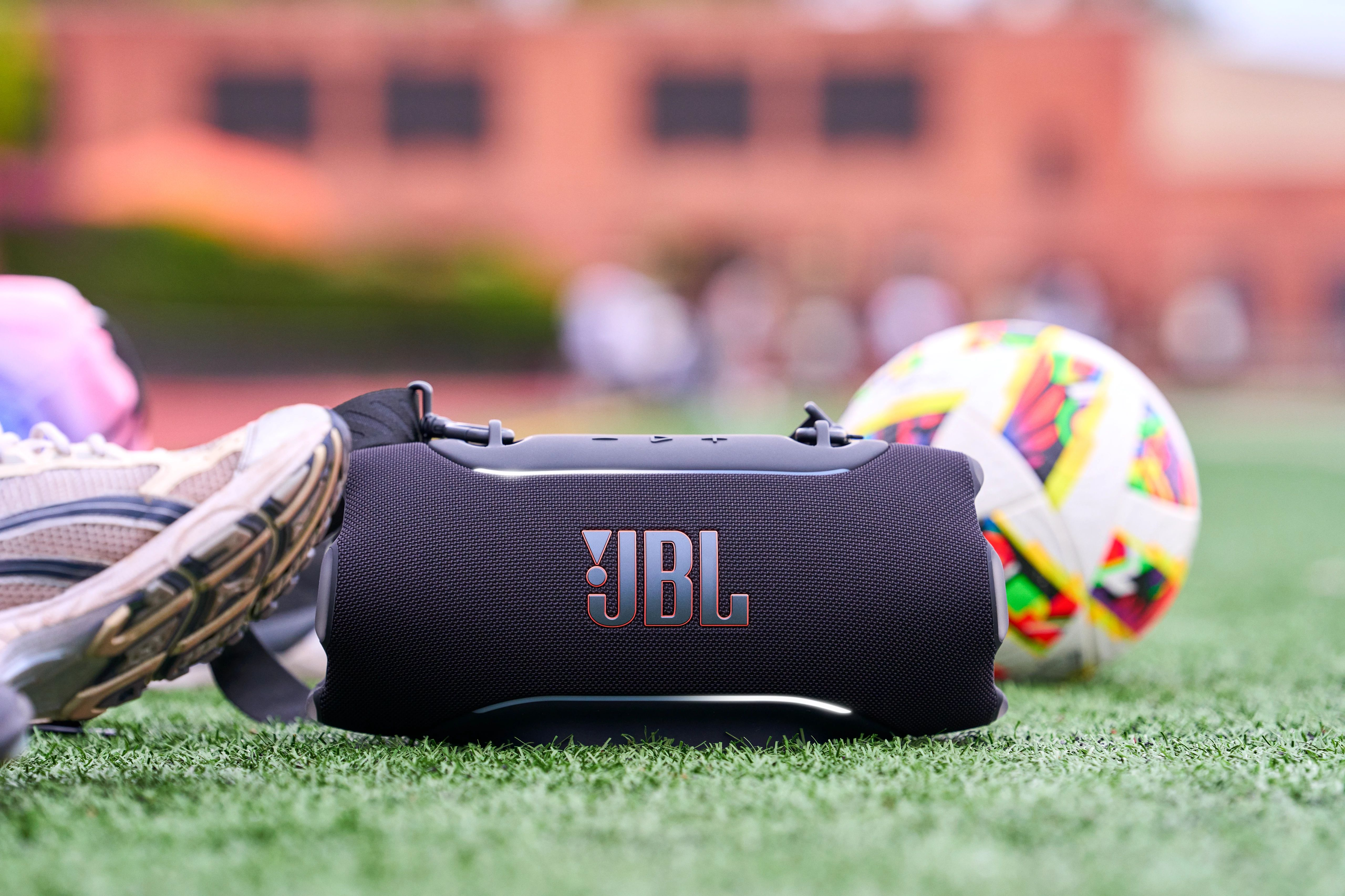 Suni çim üzerinde futbol topu ve spor ayakkabısıyla siyah JBL hoparlör.
