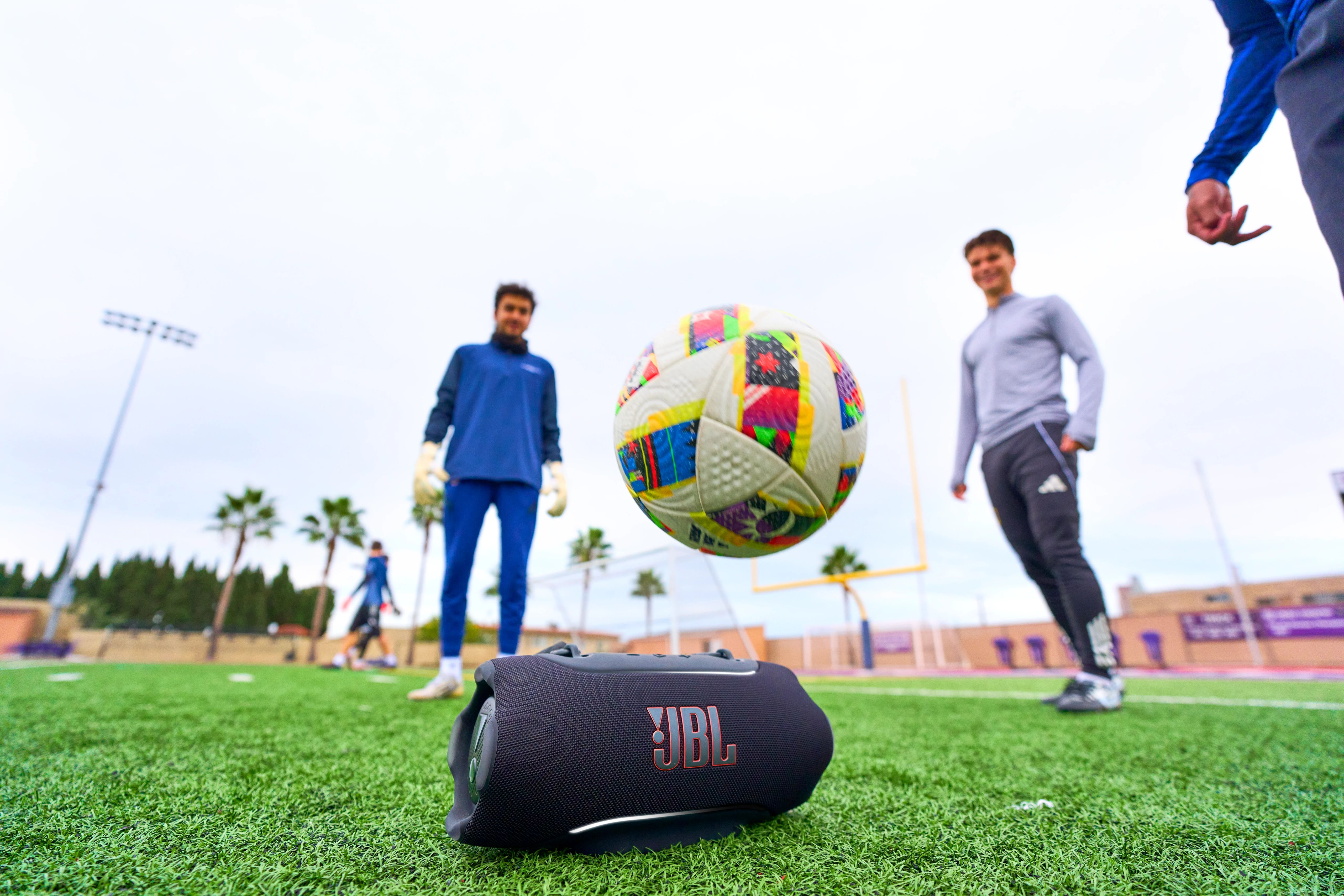 Suni çimde JBL hoparlörlü futbol topu, arka planda oyuncular.