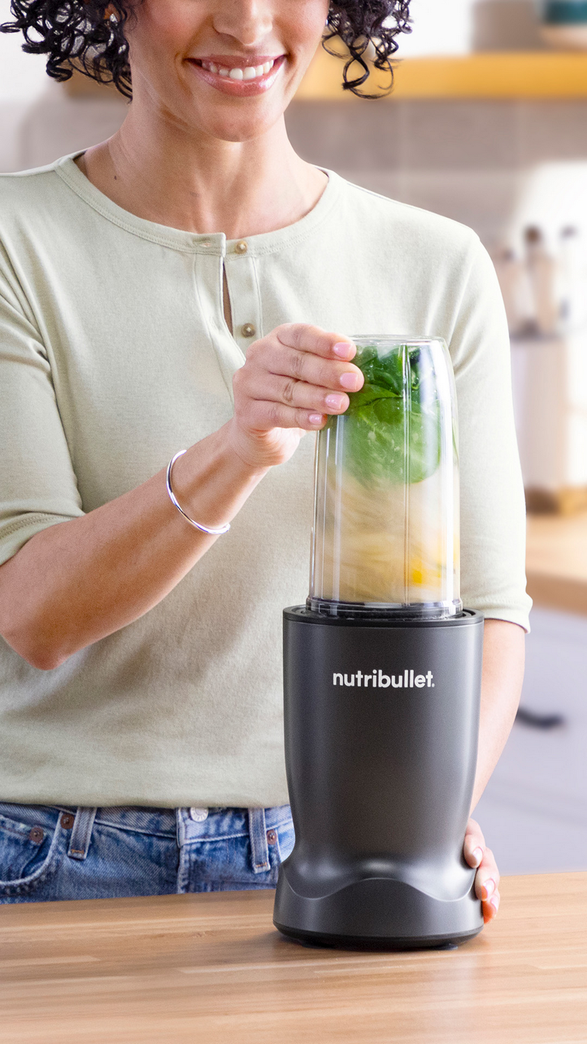 Una mujer sonriente con cabello rizado usa una licuadora Nutribullet negra con hojas verdes y trozos amarillos.