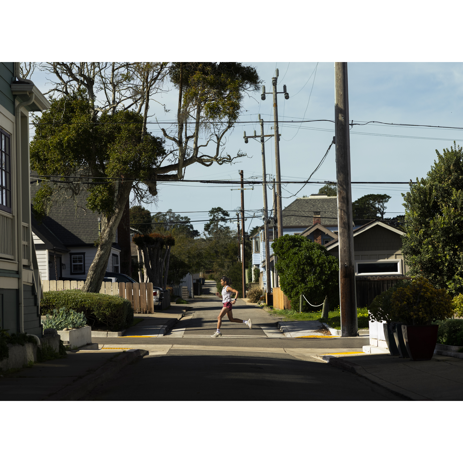 A woman runs down a residential street past houses and trees. Utility poles line the street.