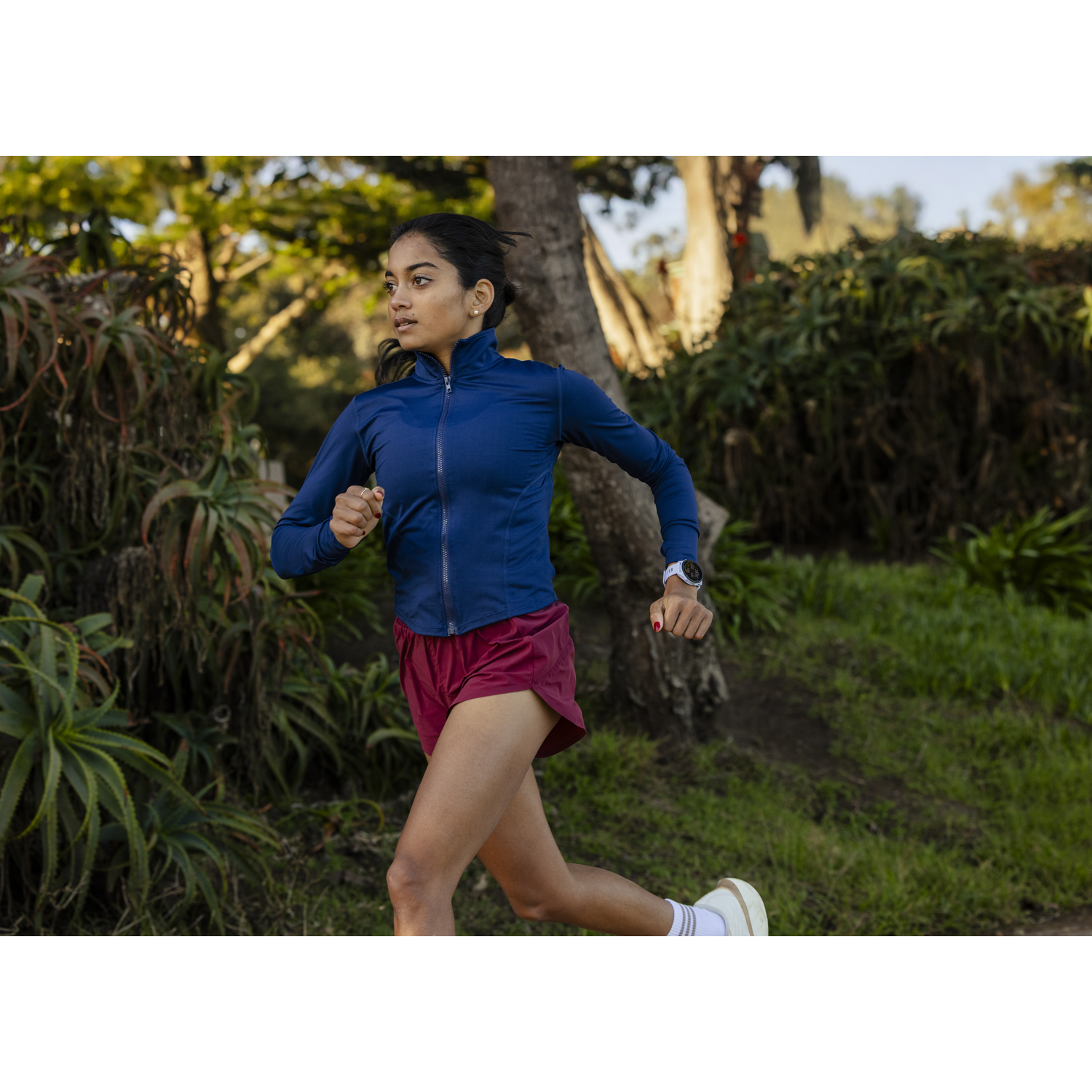 A woman in a blue jacket and maroon shorts runs outdoors, surrounded by greenery.