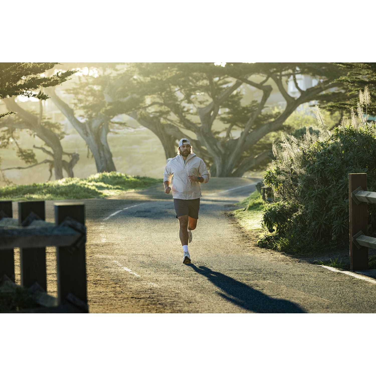 Man running on a paved path lined with trees and bushes in warm, hazy sunlight.